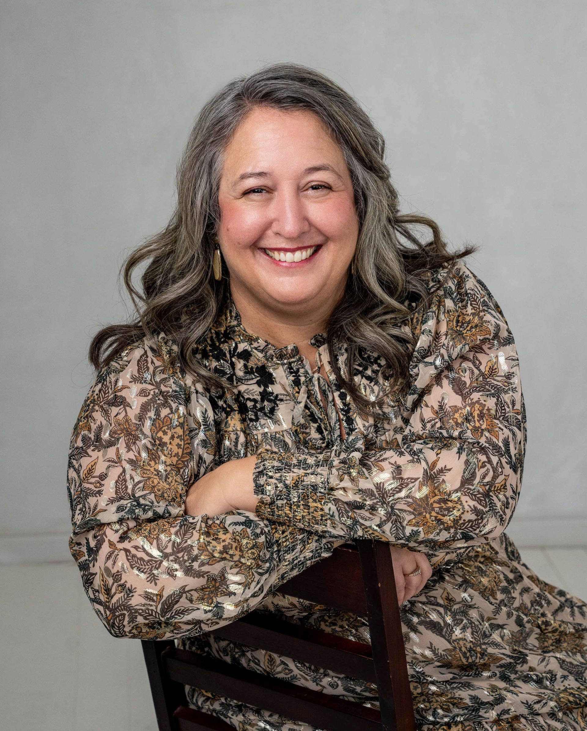 Smiling person seated on a wooden chair against a gray backdrop, wearing a patterned dress.