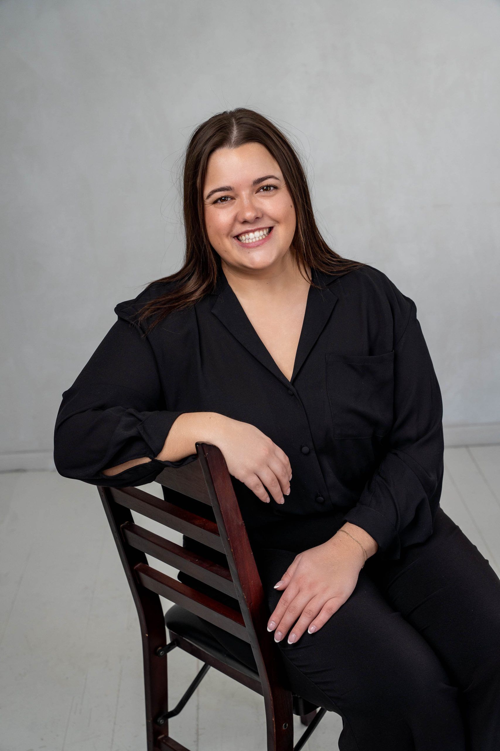 Woman in black outfit seated on a chair, smiling against a gray studio backdrop