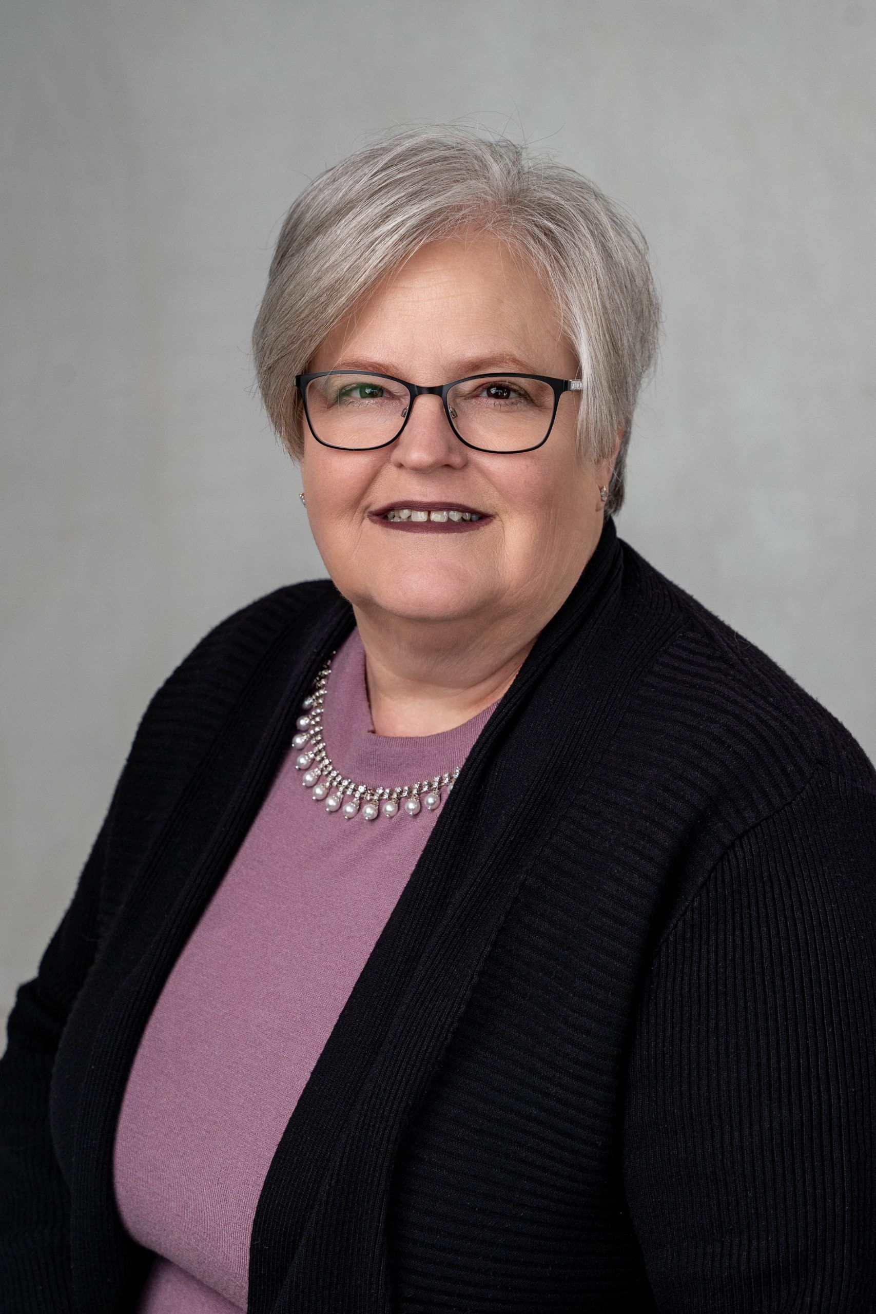 Studio portrait of a smiling person in glasses, wearing a purple top and black cardigan against a gray background