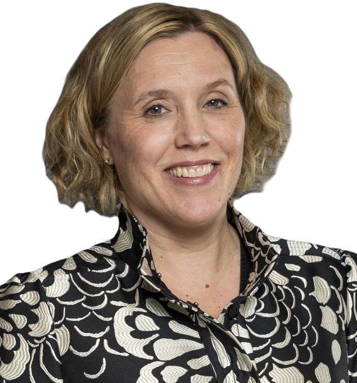 A person with shoulder-length blonde hair wearing a dark blue blazer and shirt against a plain white background.