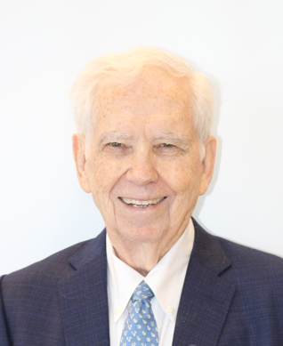 A smiling person with white hair, wearing a blue patterned tie and a navy blue suit jacket against a plain white background.