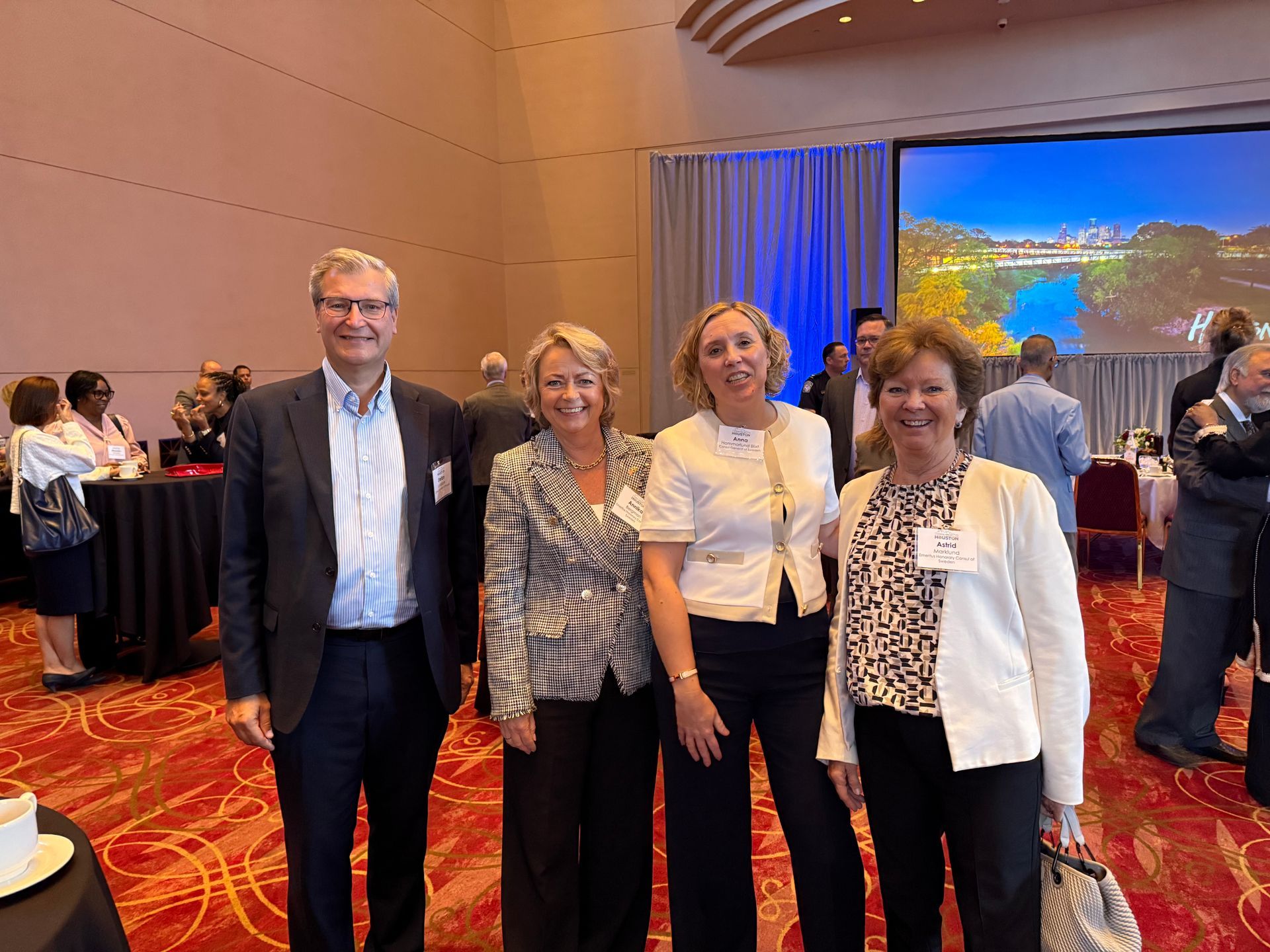 Four people pose for a photo at a banquet hall event, with a screen showing a landscape in the background.
