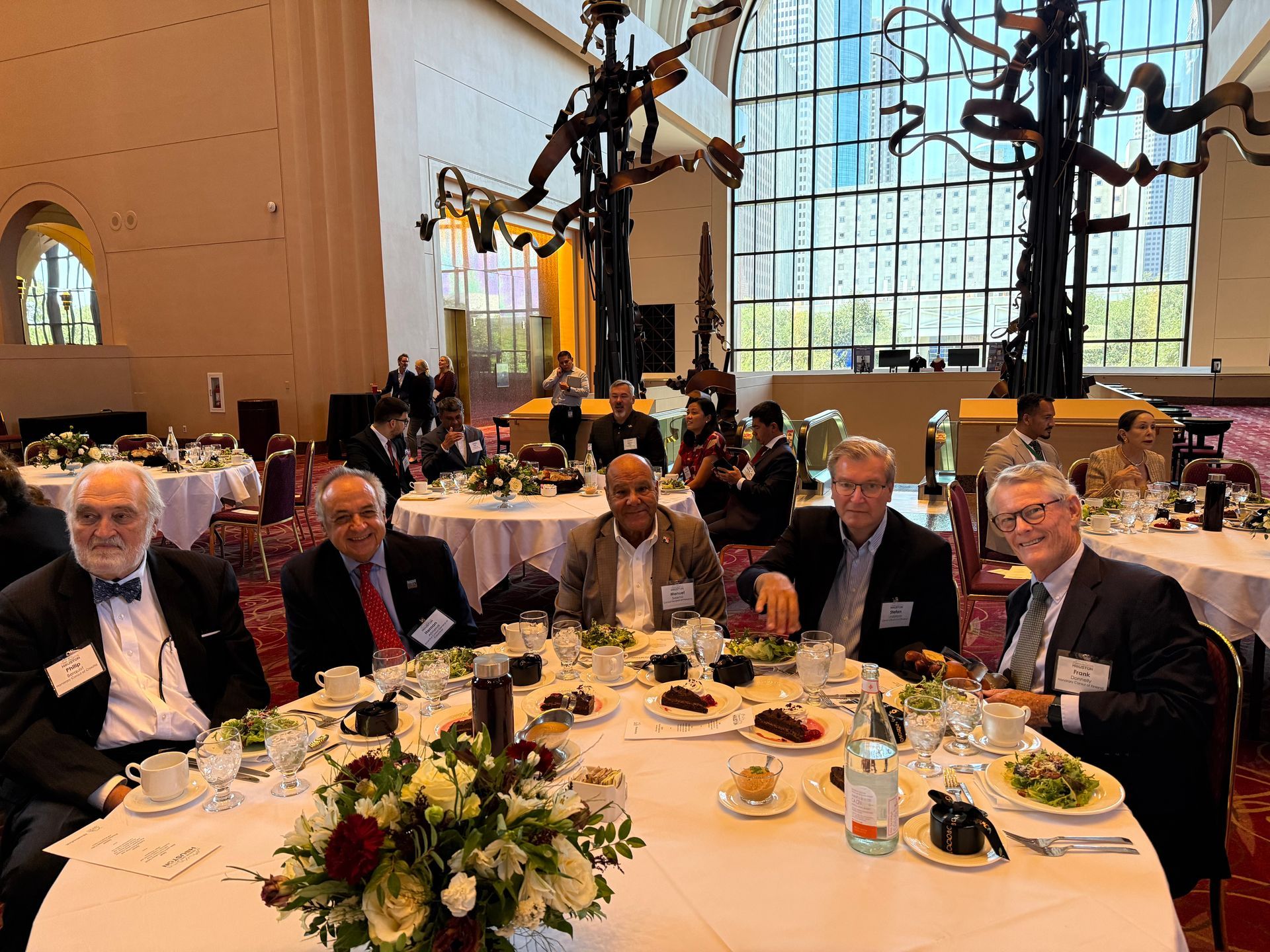Five people in suits seated at a round table for a banquet in a spacious, light-filled hall with sculptural art.