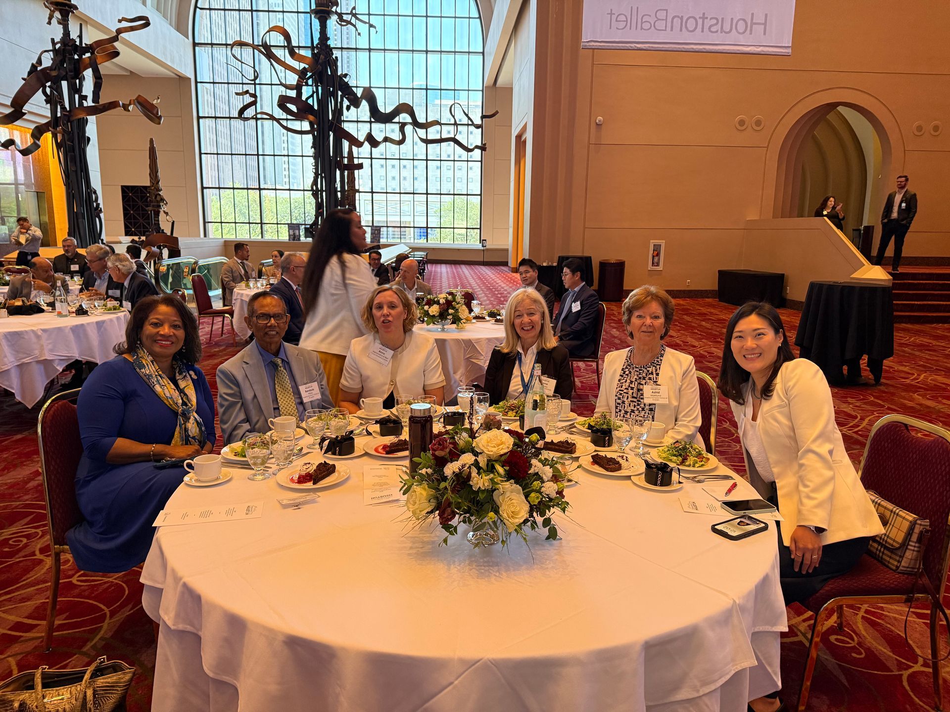 Five people sit around a round dinner table in a brightly lit, elegant atrium.