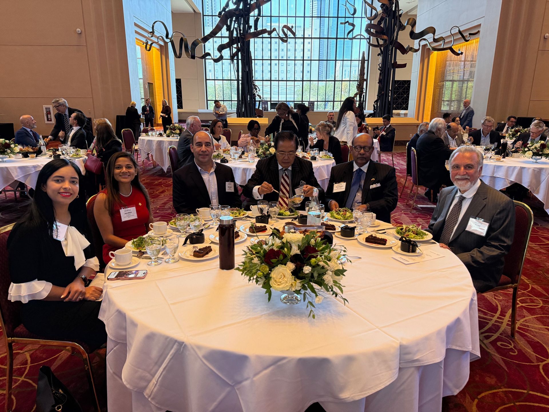 A group of people sitting at a round table at a formal event in a large, well-lit banquet hall with high ceilings.