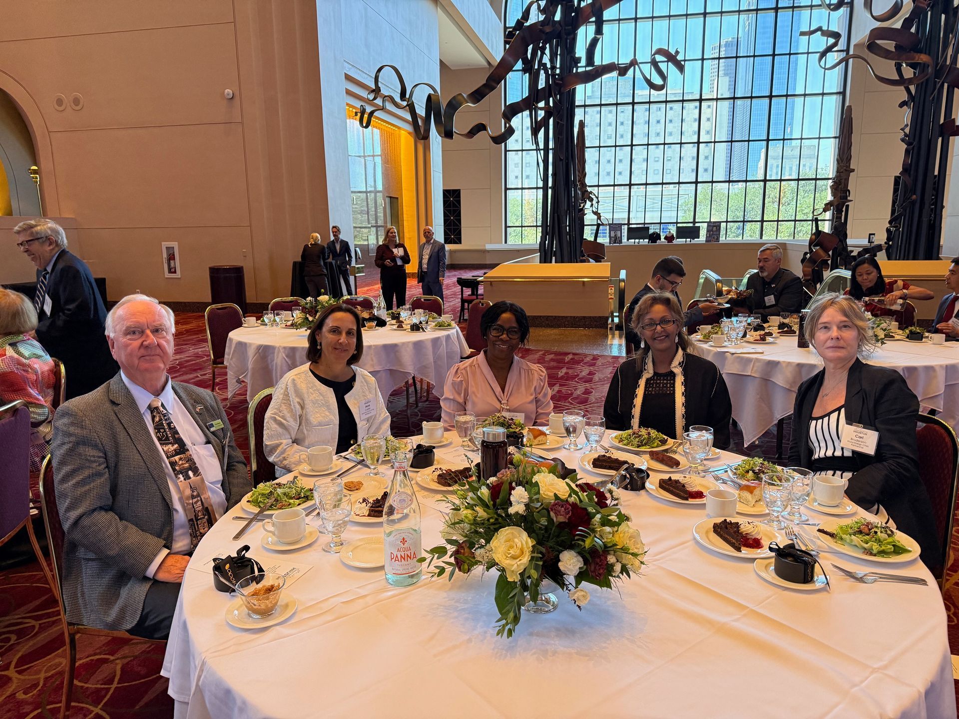 Five people sit at a round dining table in a large, modern event hall with tall windows and abstract metal sculptures.