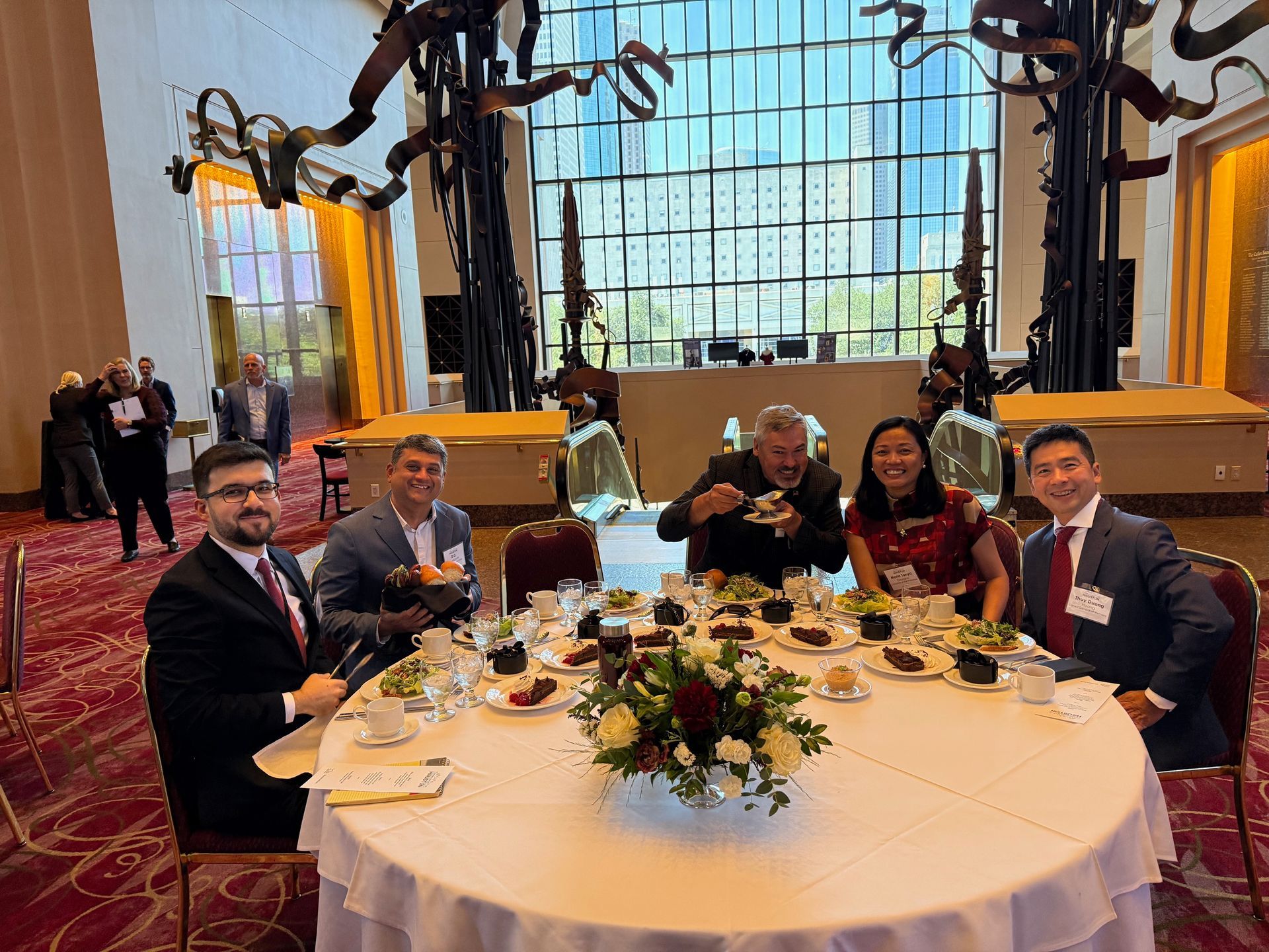 A group of people in business attire seated around a round dining table in a large, modern conference hall.