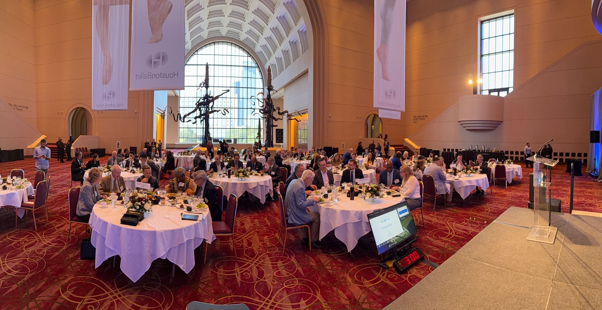 A large, banquet-style event with round tables and guests in a high-ceilinged museum hall with a red patterned carpet.