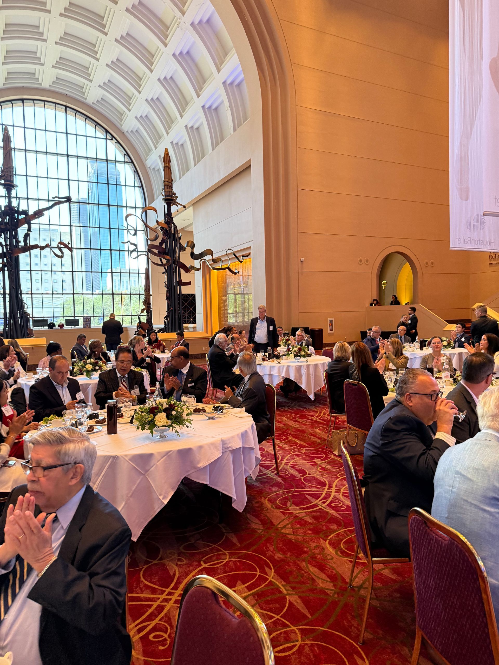 Attendees sit at round tables in a grand, high-ceilinged hall with arched windows, gathered for a formal event.