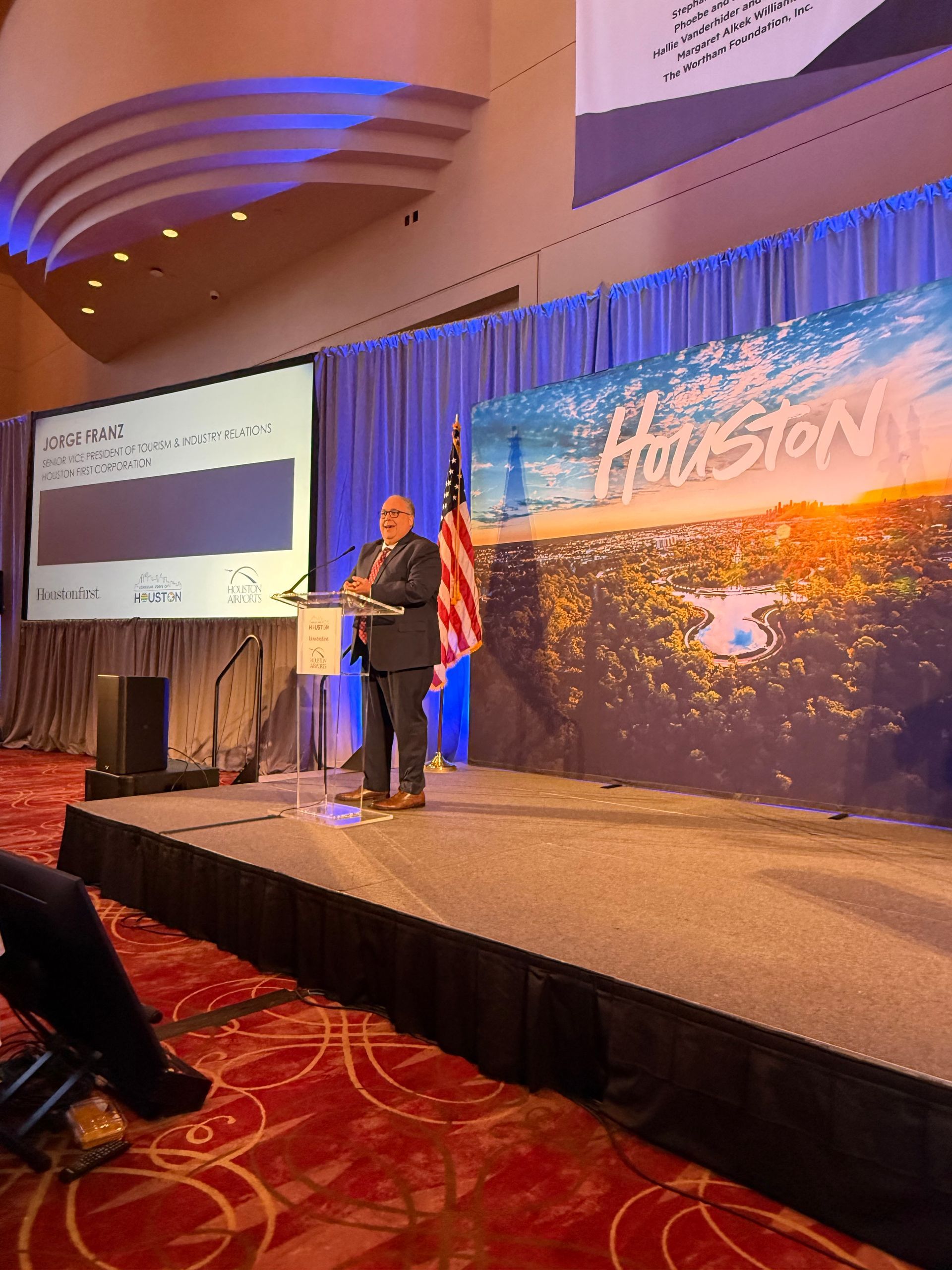 A speaker stands at a podium on a stage with a Houston skyline backdrop and an American flag in a conference hall.