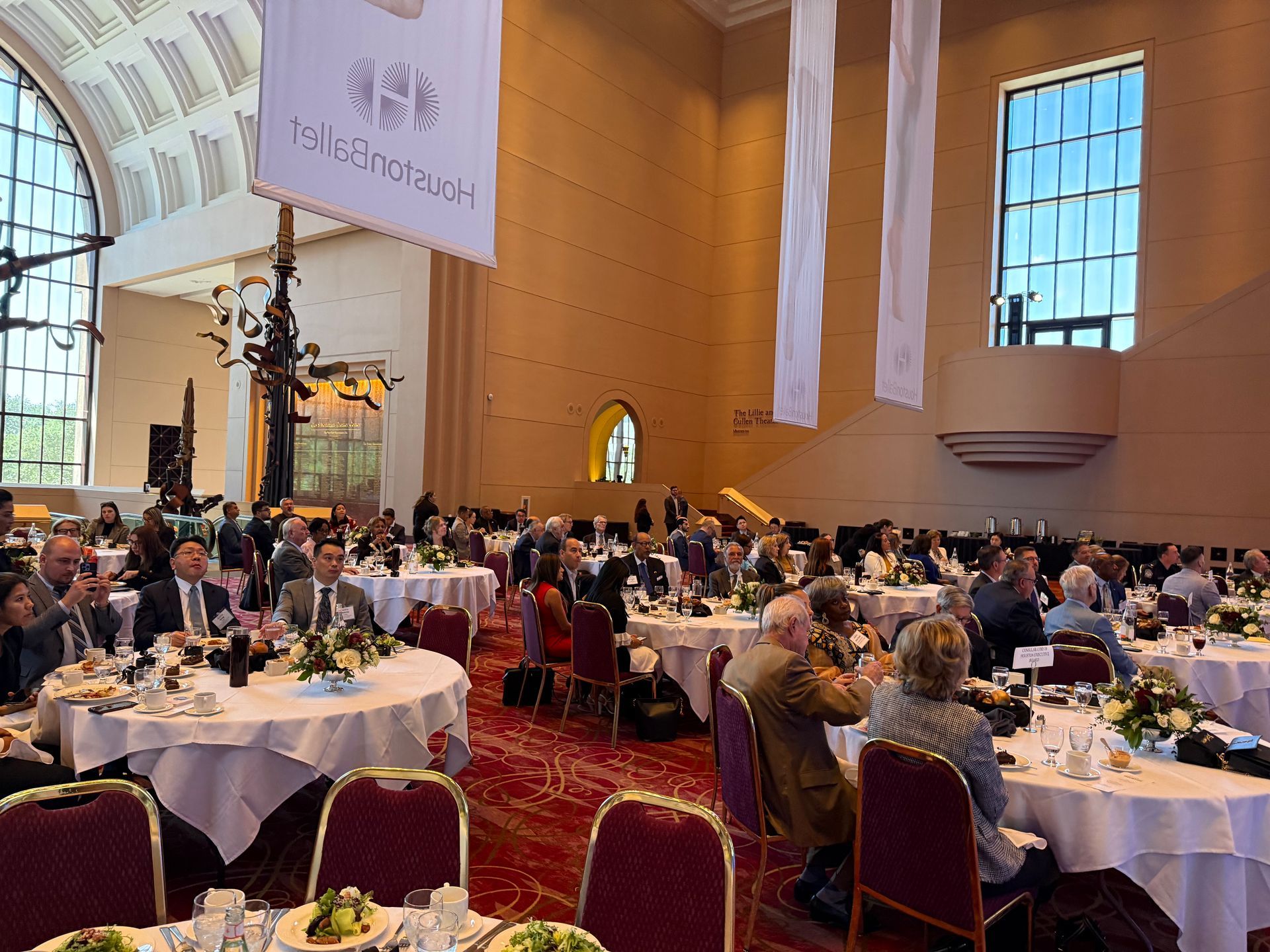 Guests dine at round tables in a large, high-ceilinged room with tall windows and a patterned red carpet.