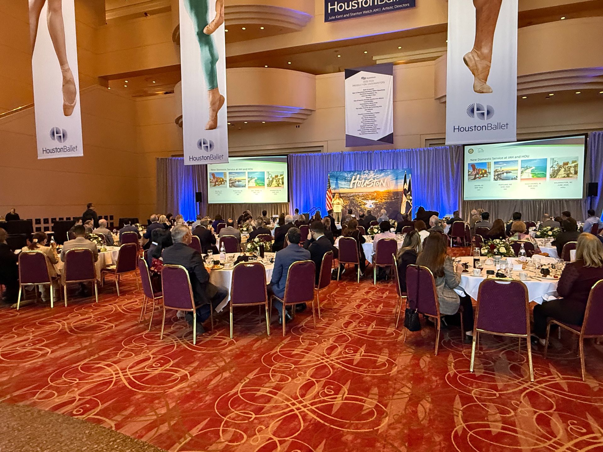 A conference ballroom with guests seated at round tables facing a stage with two large projection screens and banners.