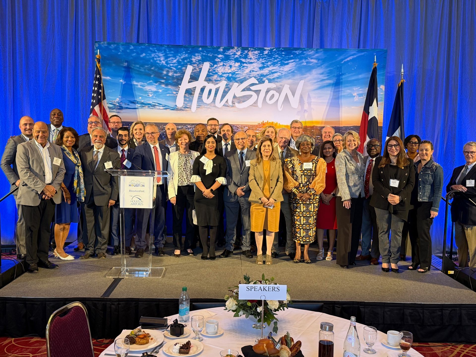 A large group of people stands on a stage in front of a blue backdrop with the word