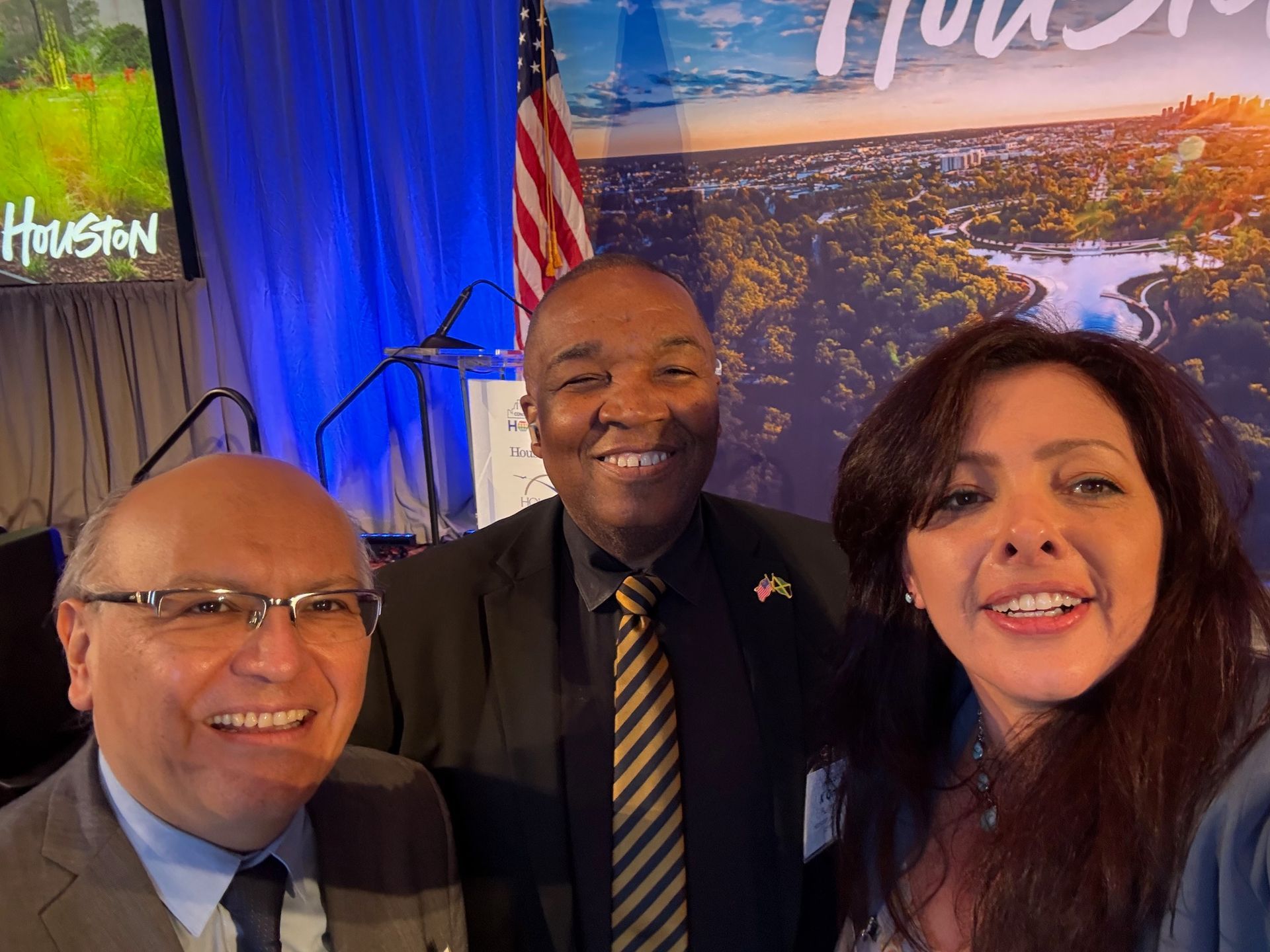 Three people smiling for a selfie at an event featuring a Houston-themed backdrop and an American flag.