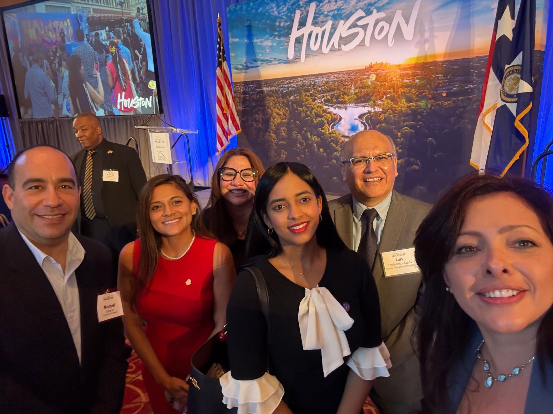 Six people pose for a selfie at a Houston-themed event in front of a city backdrop and American and Texas flags.