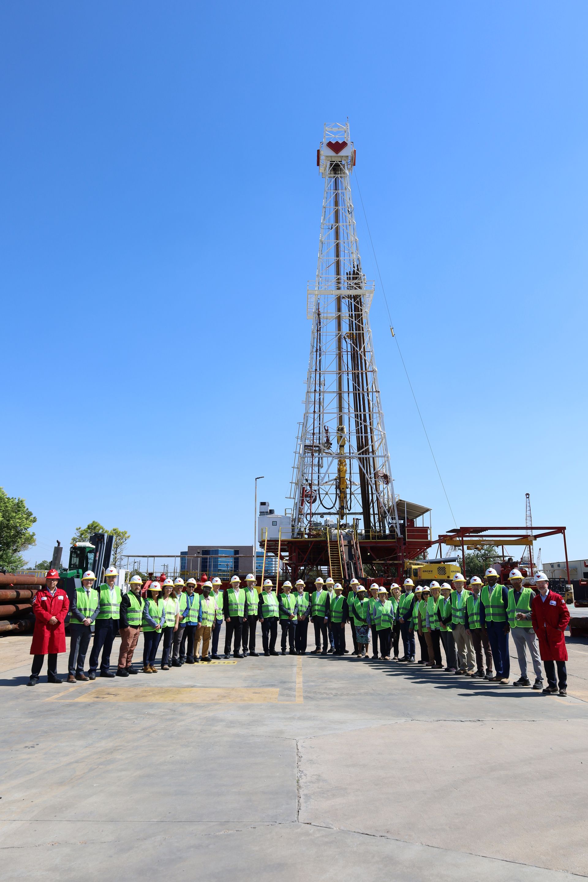 Group in green shirts posing before a tall drilling rig under a clear blue sky.