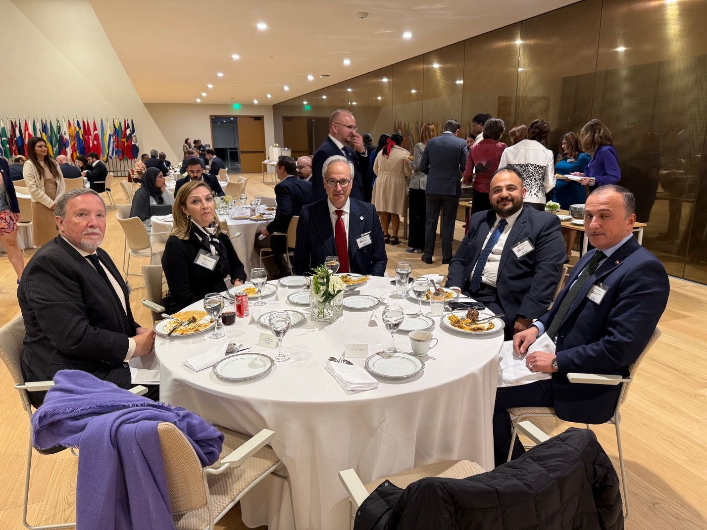 Five professionals sit at a round table during a formal banquet, with other guests mingling in a brightly lit hall.