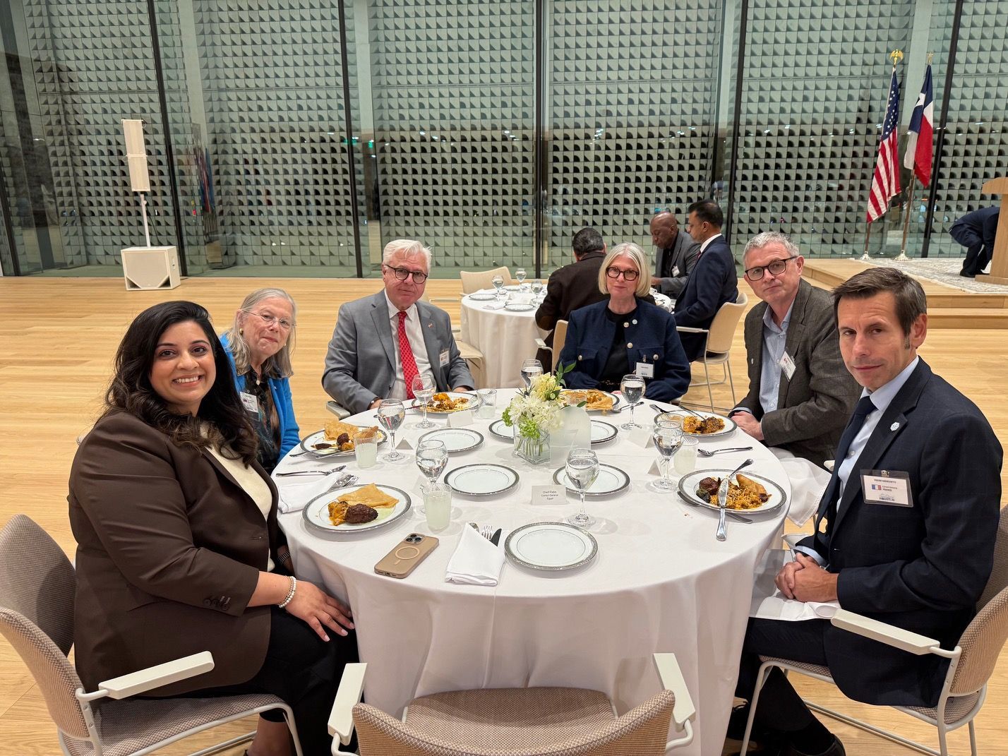 Six people sit around a round dining table in a large, modern hall with a grid-patterned glass wall.