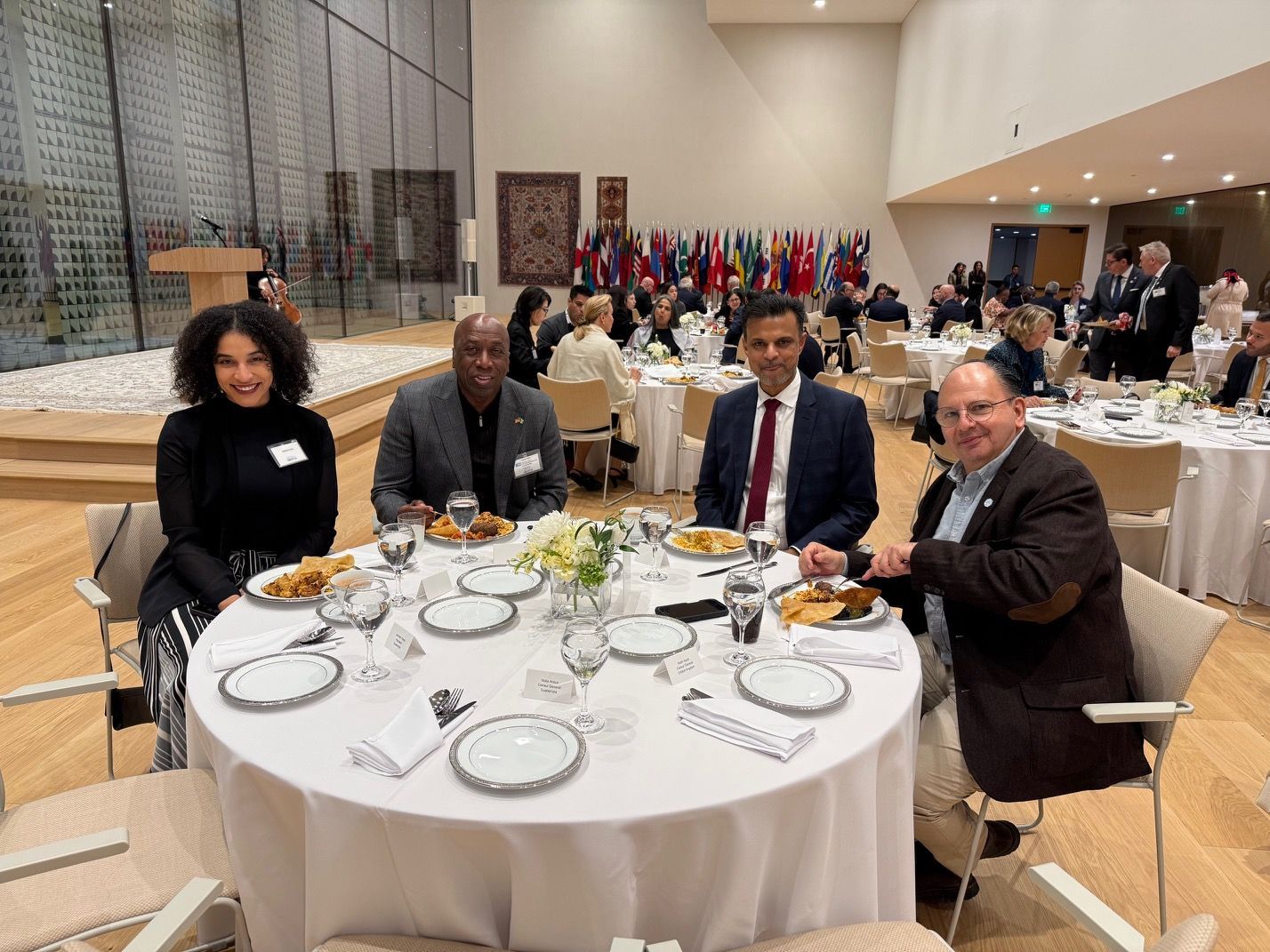 Four people sit at a round table having lunch in a large hall with international flags in the background.