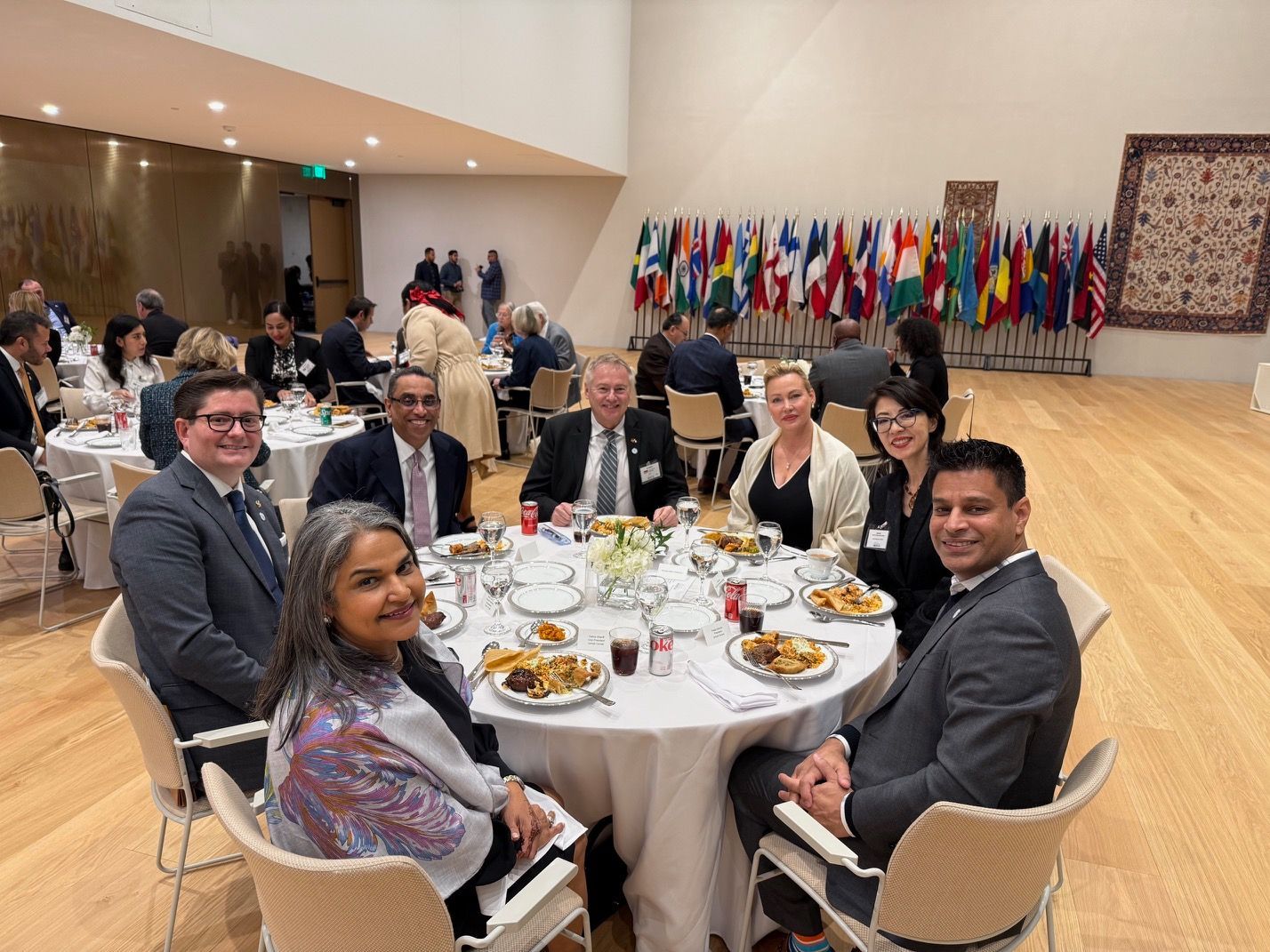 A group of people sitting at a round table during a formal banquet with a row of international flags in the background.