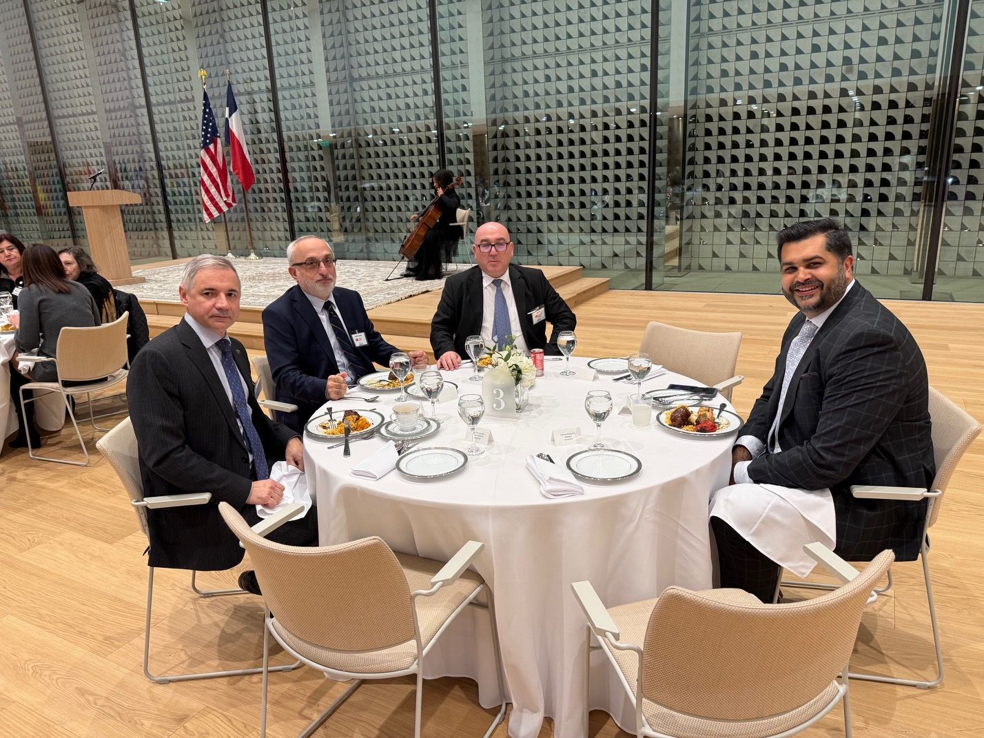 Four men in suits sit at a round table at a formal event with flags in the background.