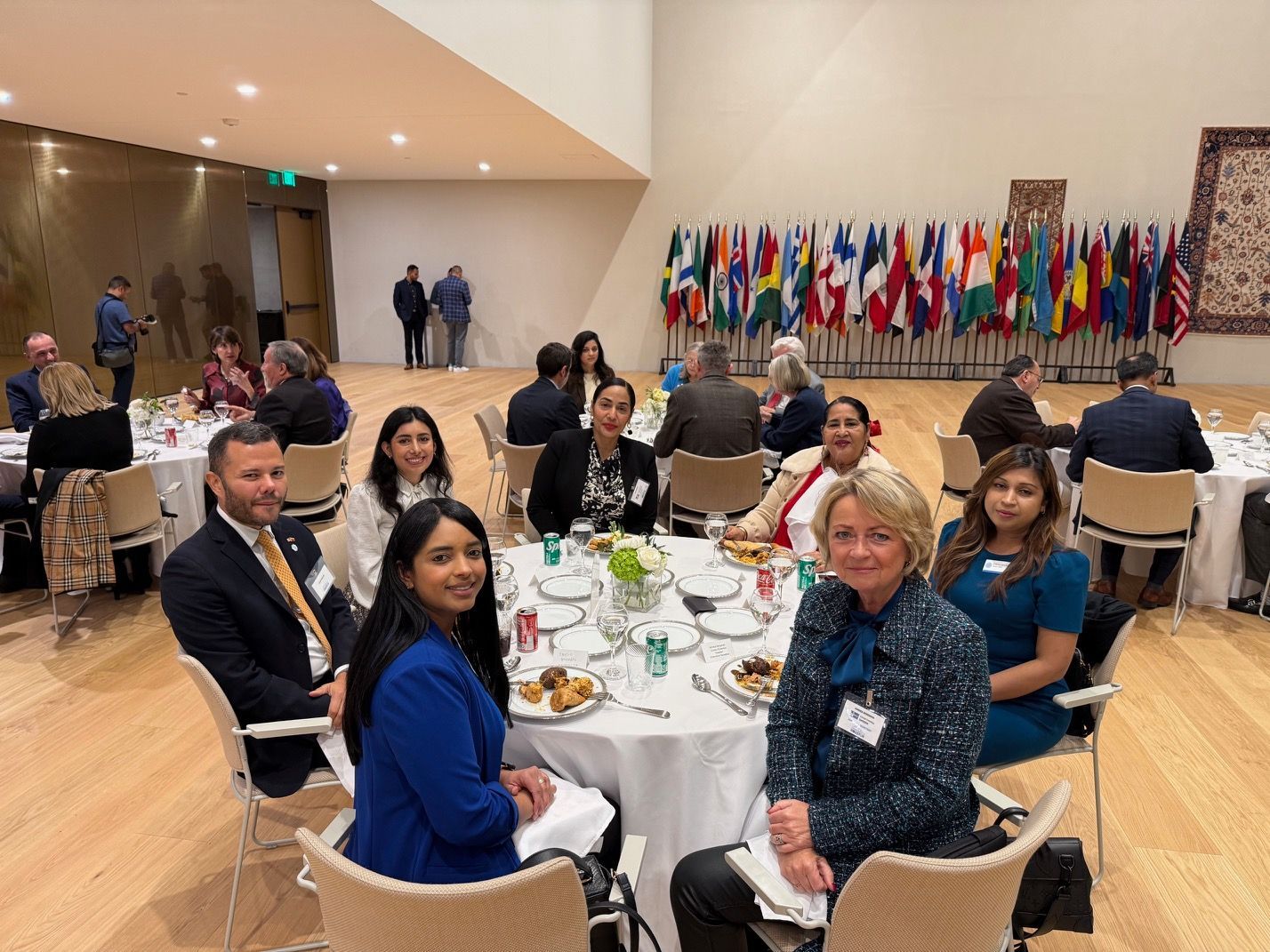 Attendees at a formal event sit at a round table for a meal in a room decorated with a row of international flags.
