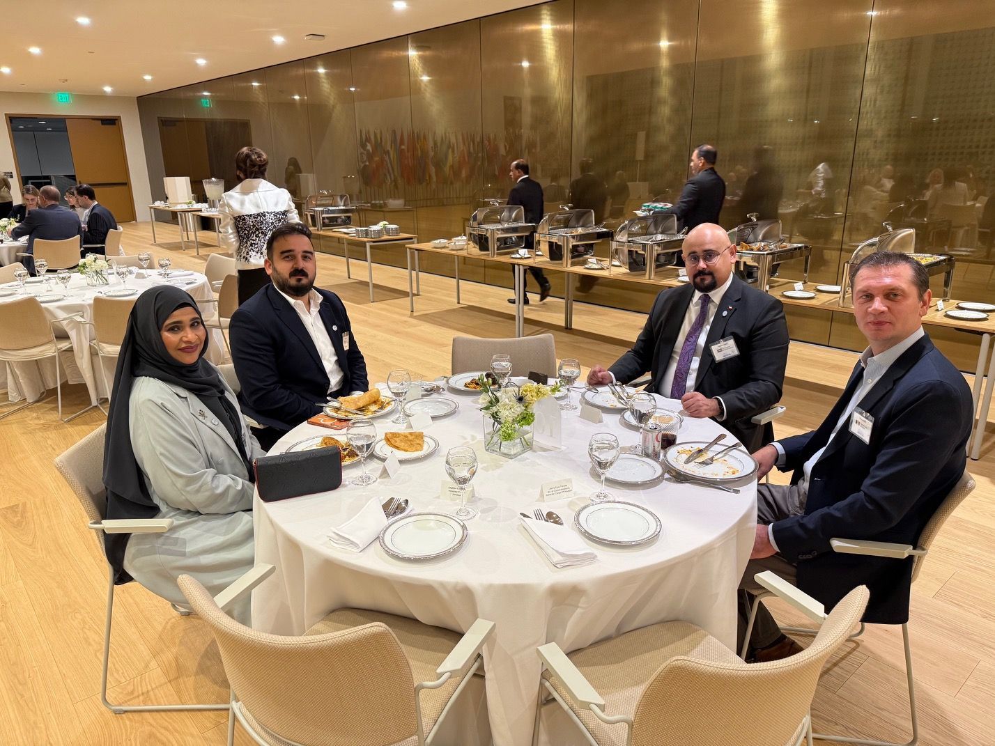 Four people in professional attire seated around a round dining table in a well-lit event hall with buffet service.