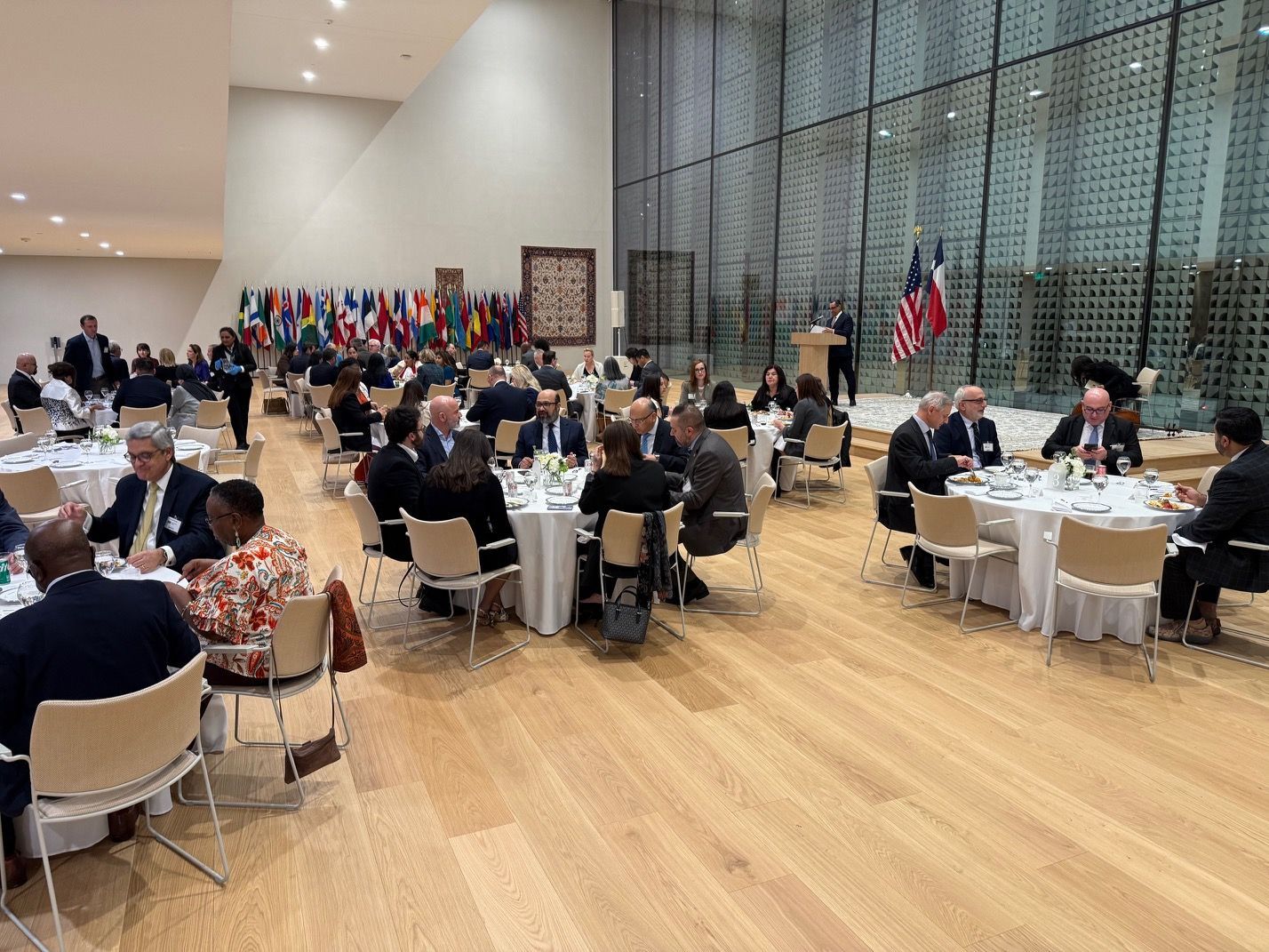 A formal banquet or conference setting with round tables, attendees, and flags in a large hall with a glass wall.