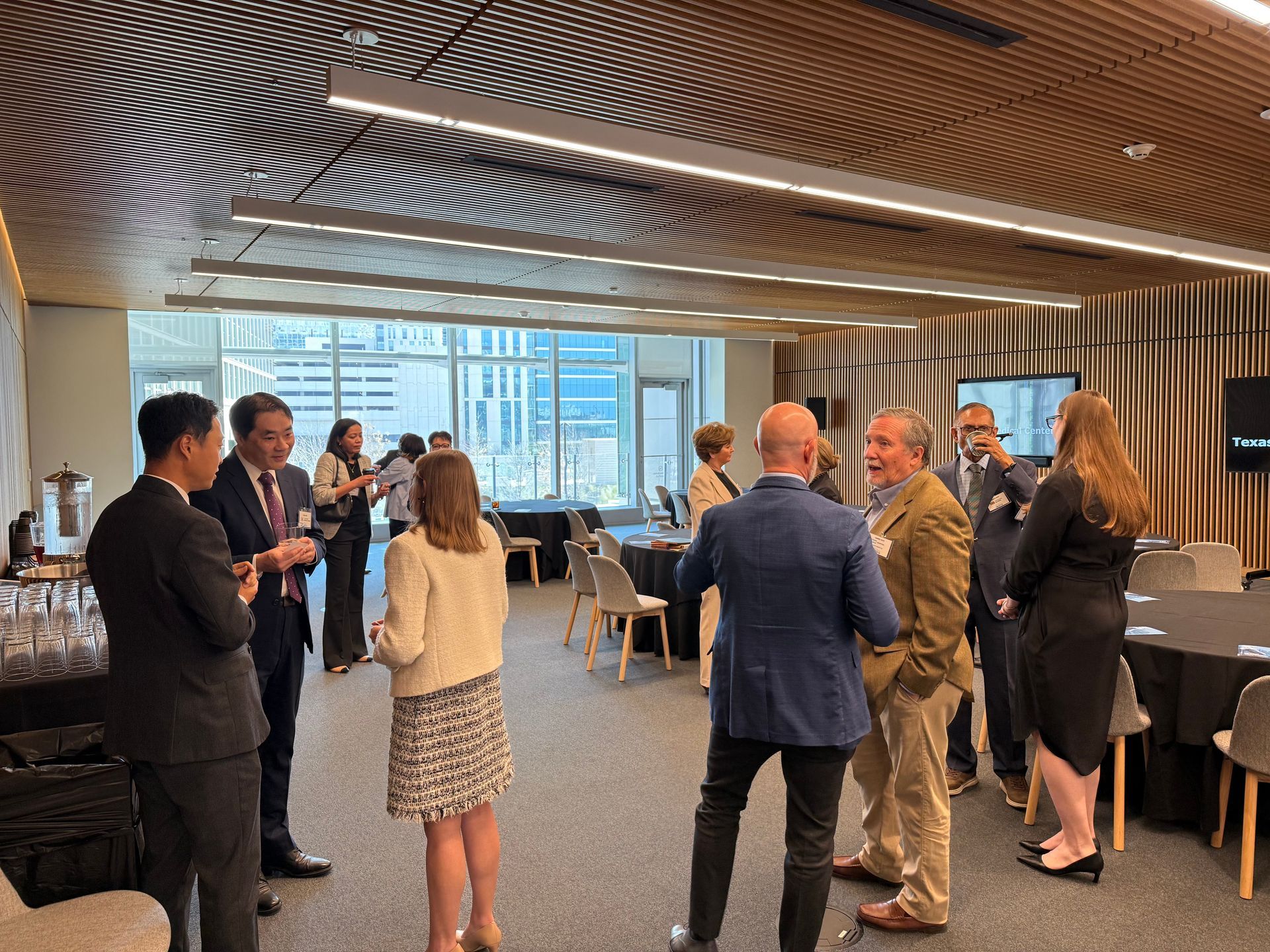 People networking at an indoor professional event with floor-to-ceiling windows and a textured ceiling.