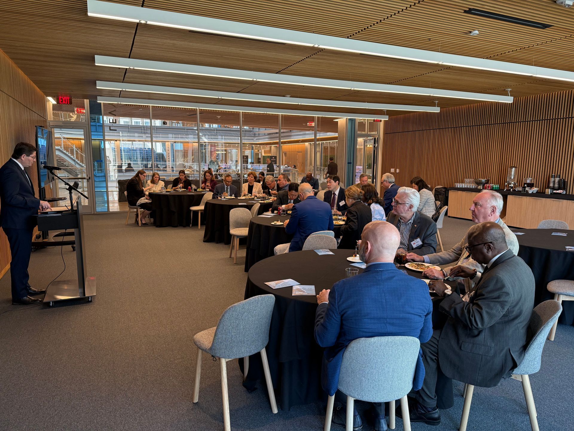 A speaker stands at a podium addressing an audience seated at round tables in a modern, well-lit conference room.