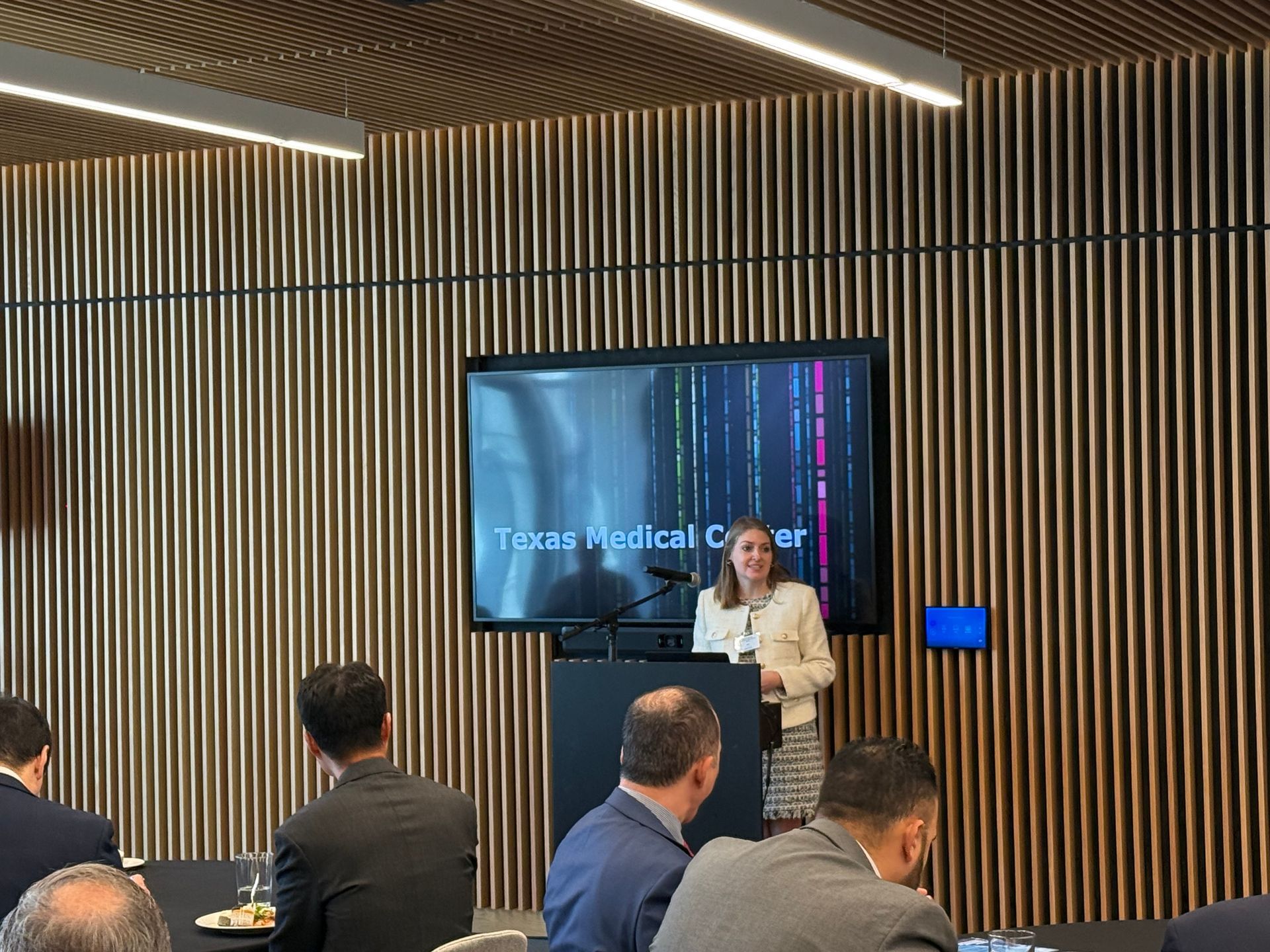 A woman presents at a podium in front of a digital screen labeled