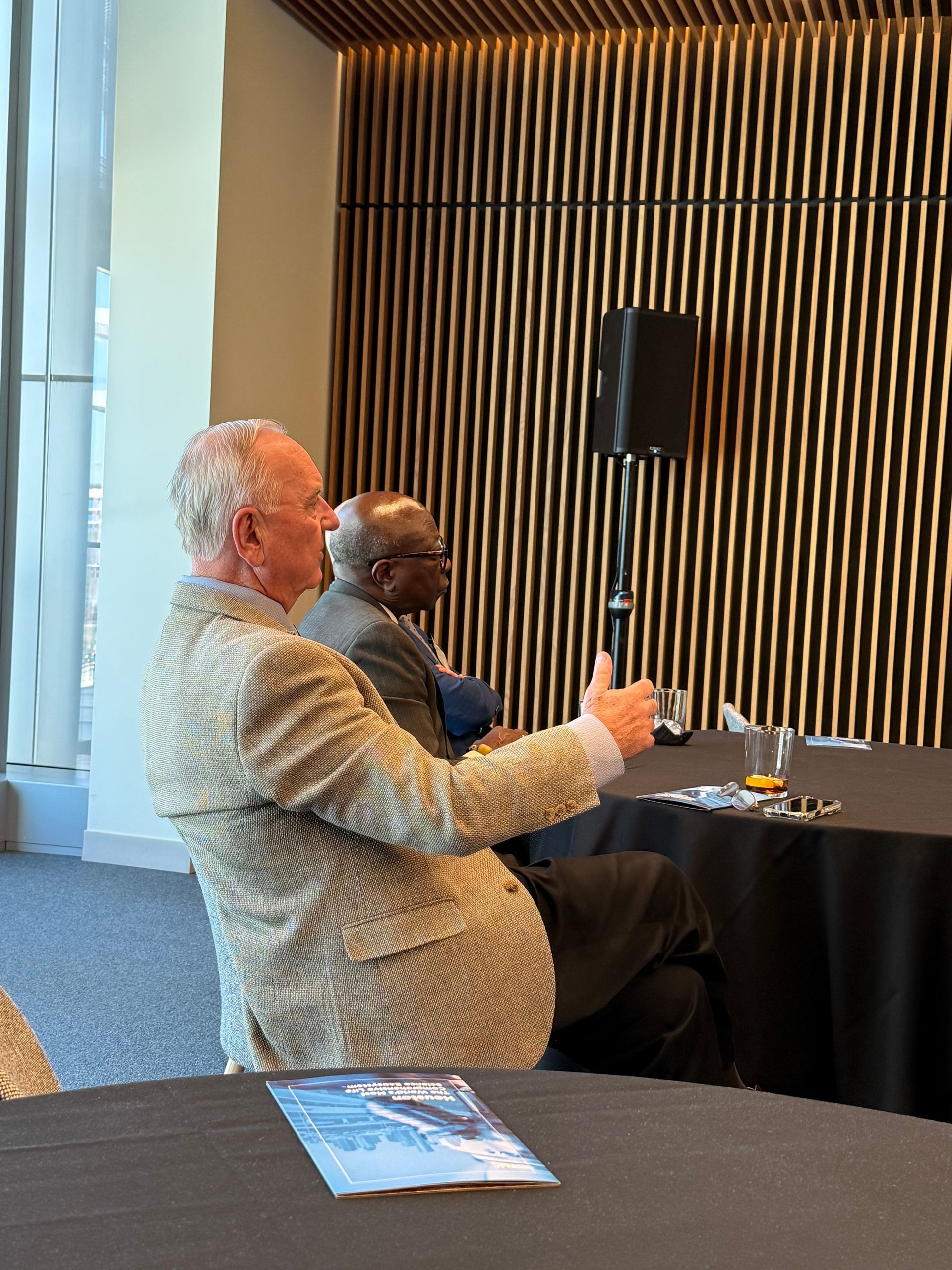 Two people sitting at round tables in a conference room with wood-paneled walls, one gesturing during a conversation.