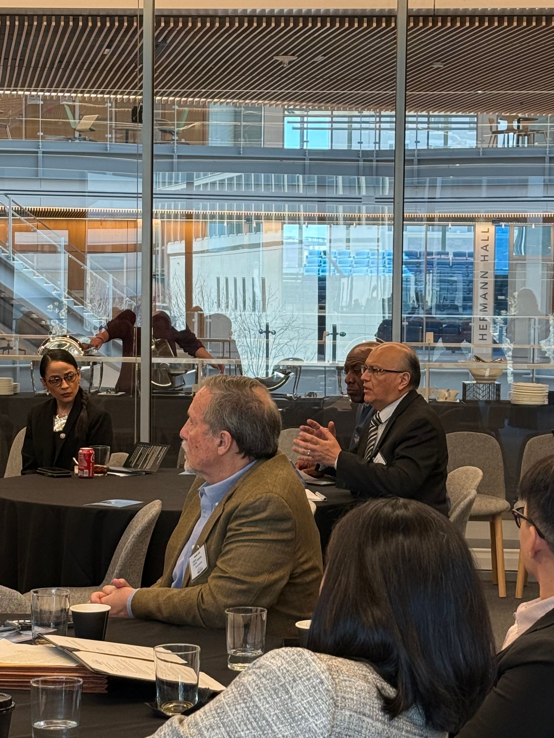 People seated at round tables during a professional event in a modern, glass-walled conference room.