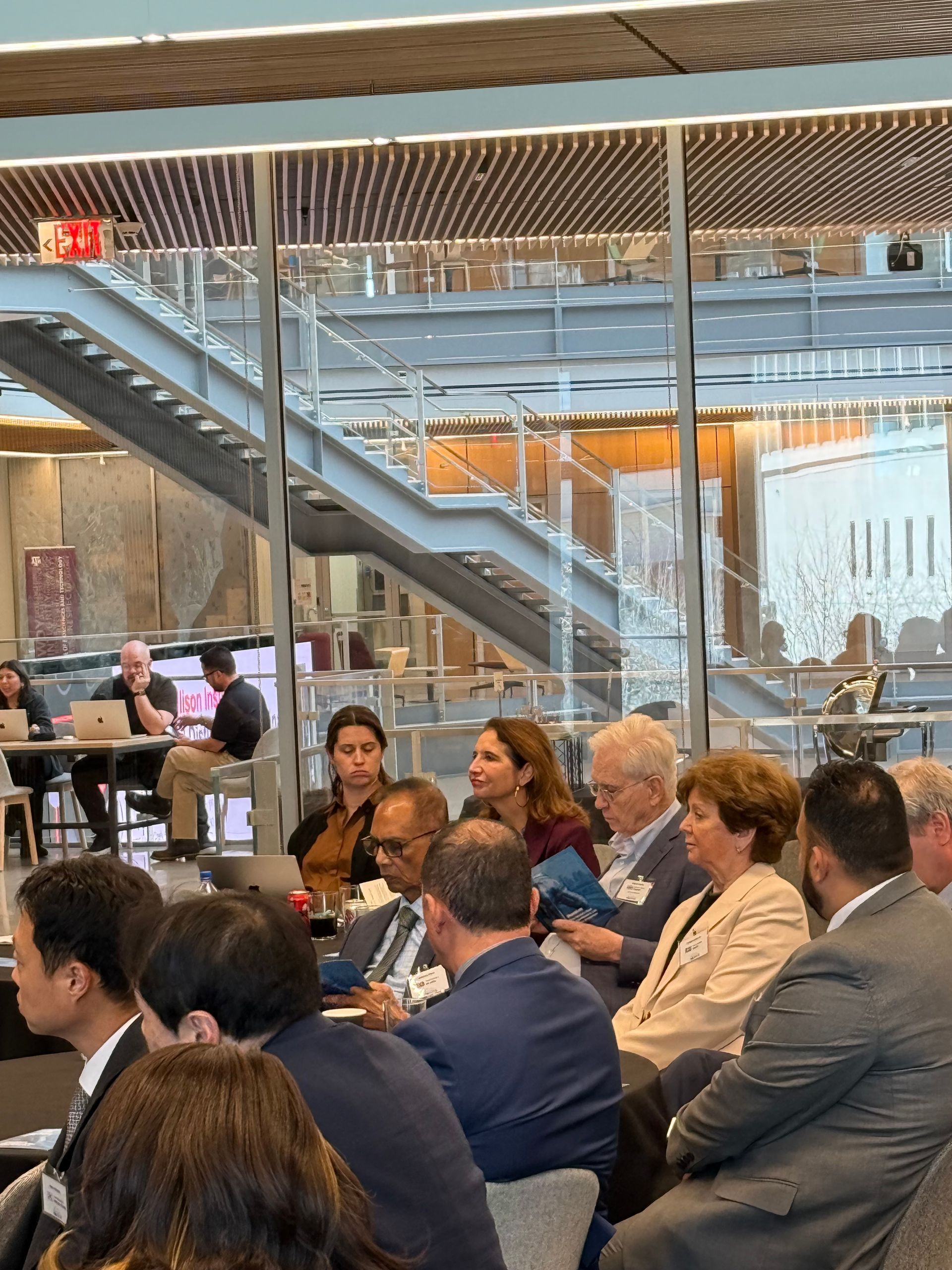 A seated audience listens to a presentation inside a modern building with glass walls and a prominent staircase.