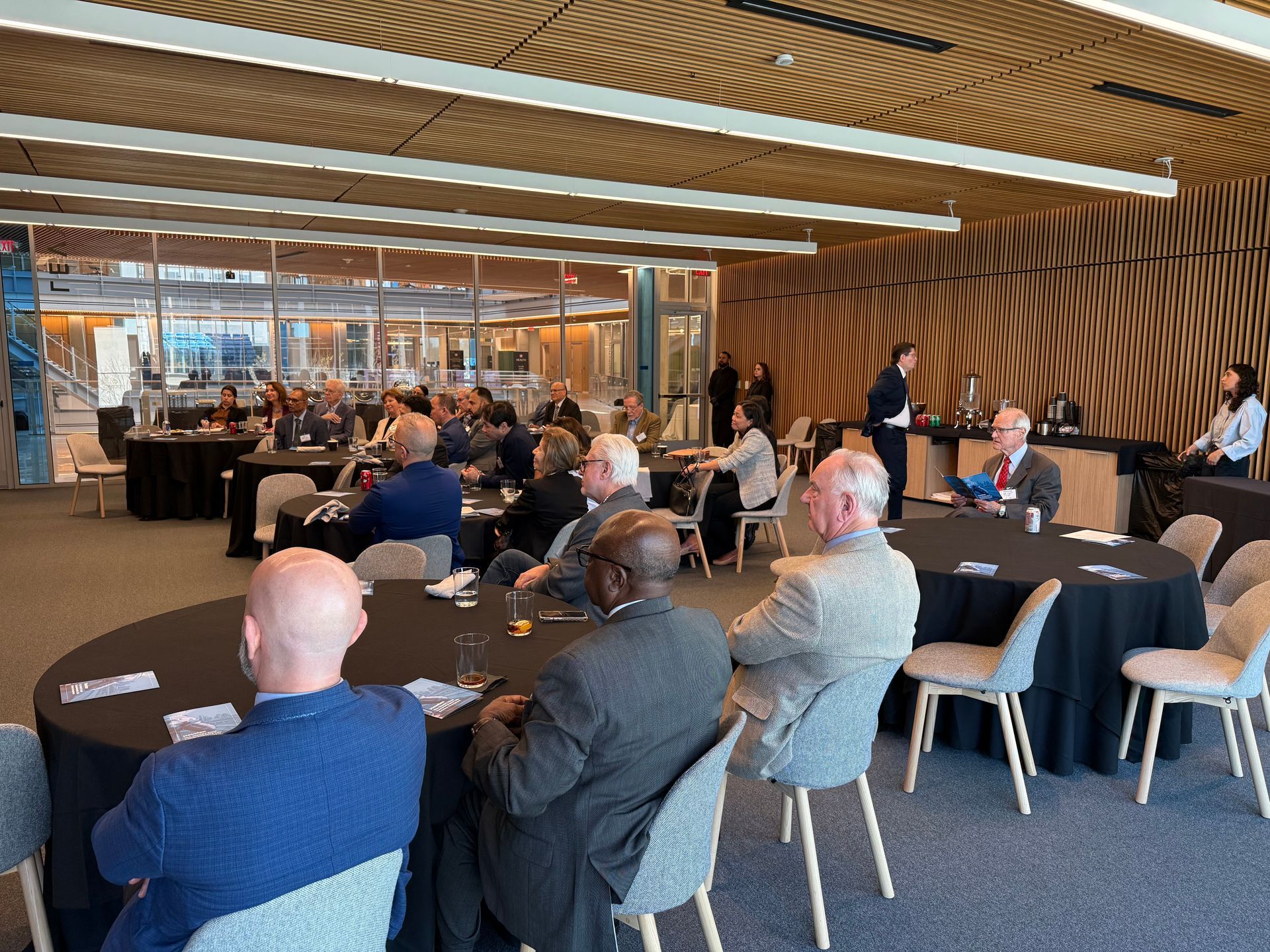 Attendees sit at circular tables with black tablecloths in a modern event space with wood-paneled walls and ceilings.