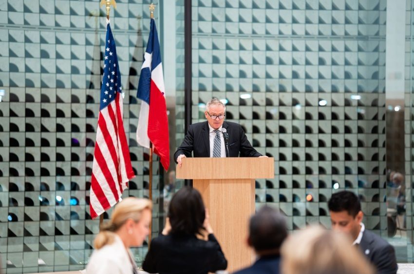 A man in a suit speaks from a wooden podium in front of the U.S. and Texas flags, with an audience in the foreground.