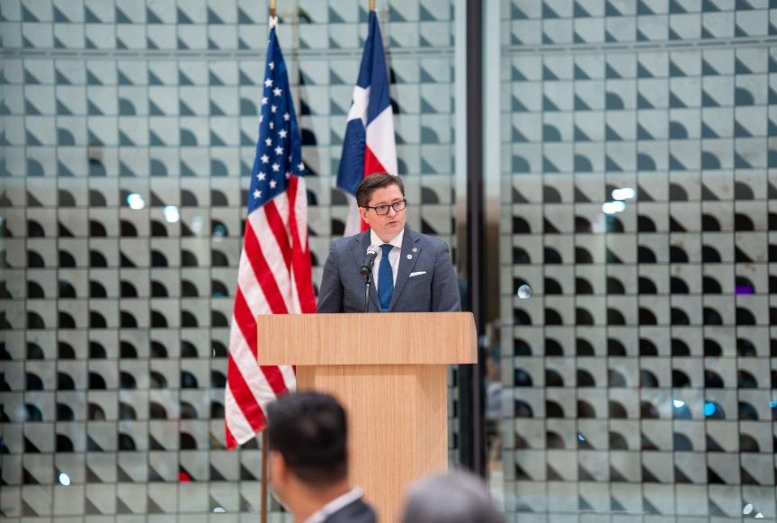 A person in a suit stands behind a podium with the U.S. and Dominican Republic flags in a modern room with a grid wall.