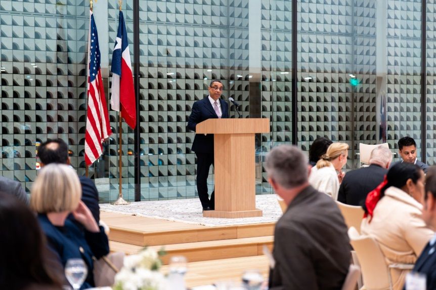 A person in a suit speaks at a wooden podium before an audience, with the U.S. and Texas flags standing in the background.