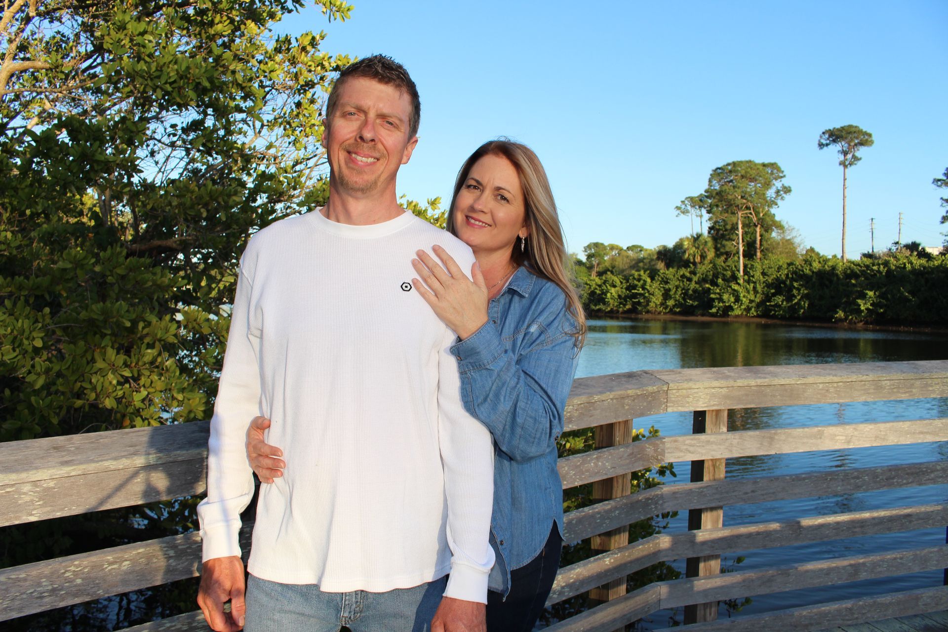 Couple standing on a wooden bridge by a river, smiling in the sunlight.