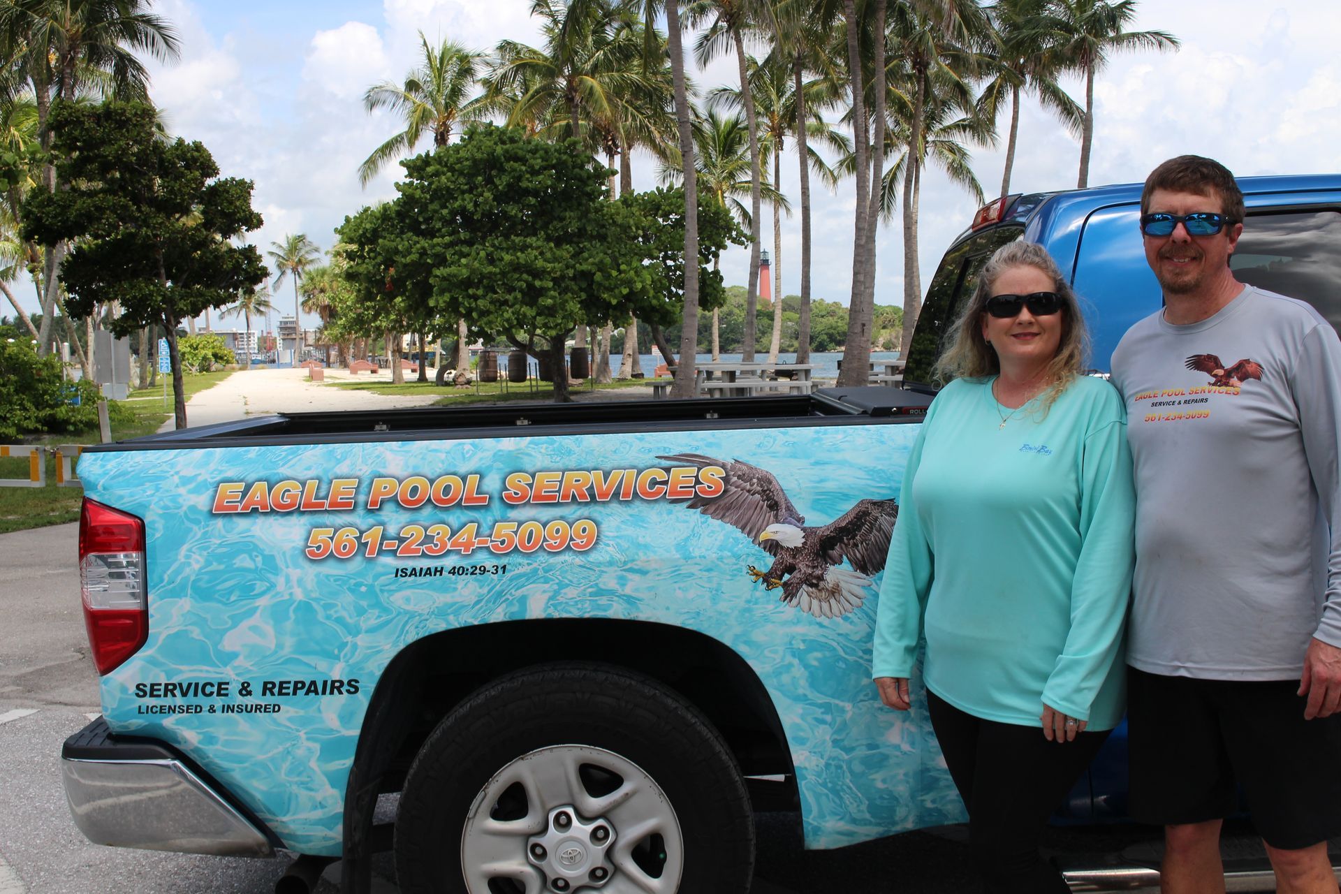 Two people posing beside a blue truck with tropical palm trees in the background.