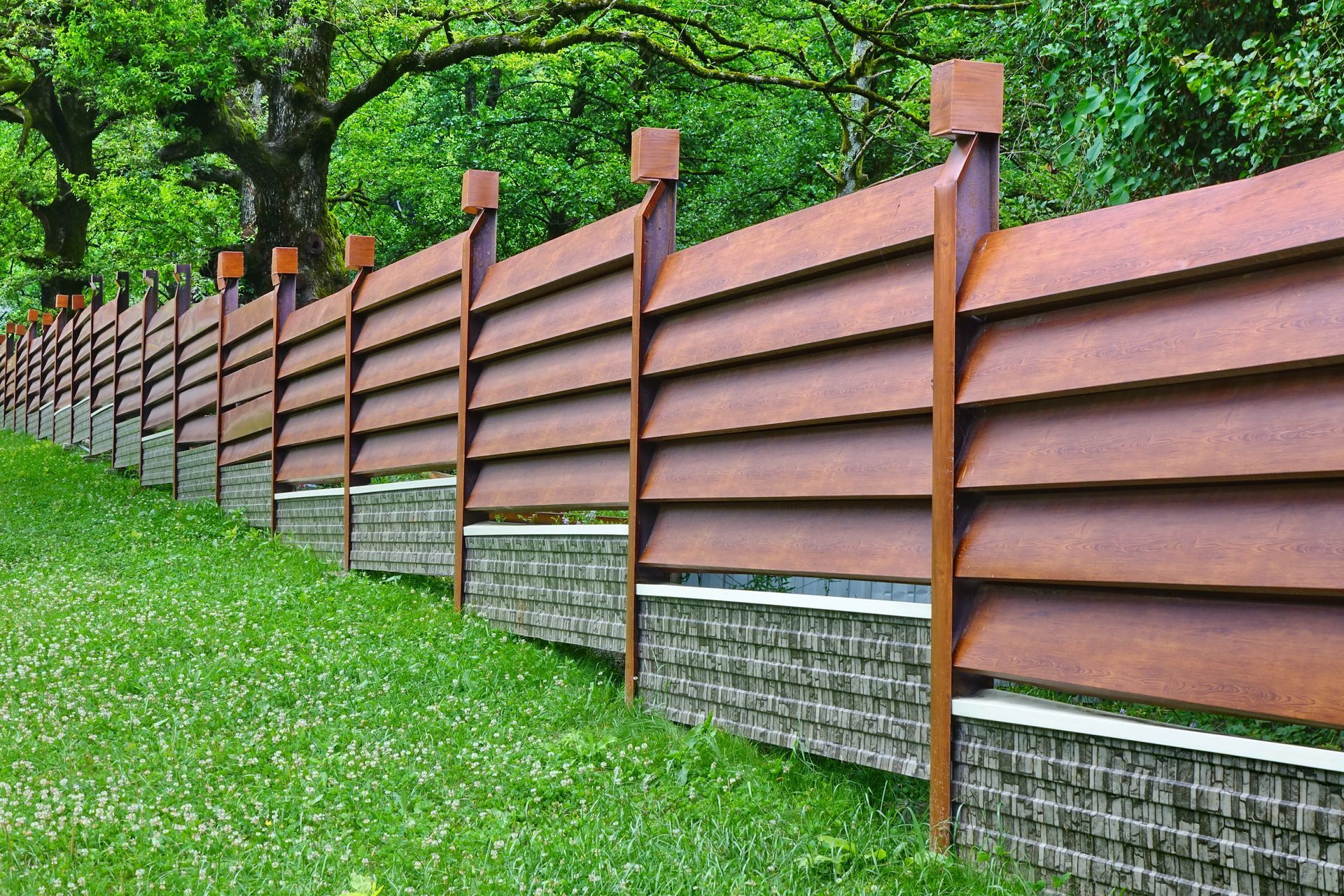 Brown slatted fence with stone base in a grassy yard, trees in the background.