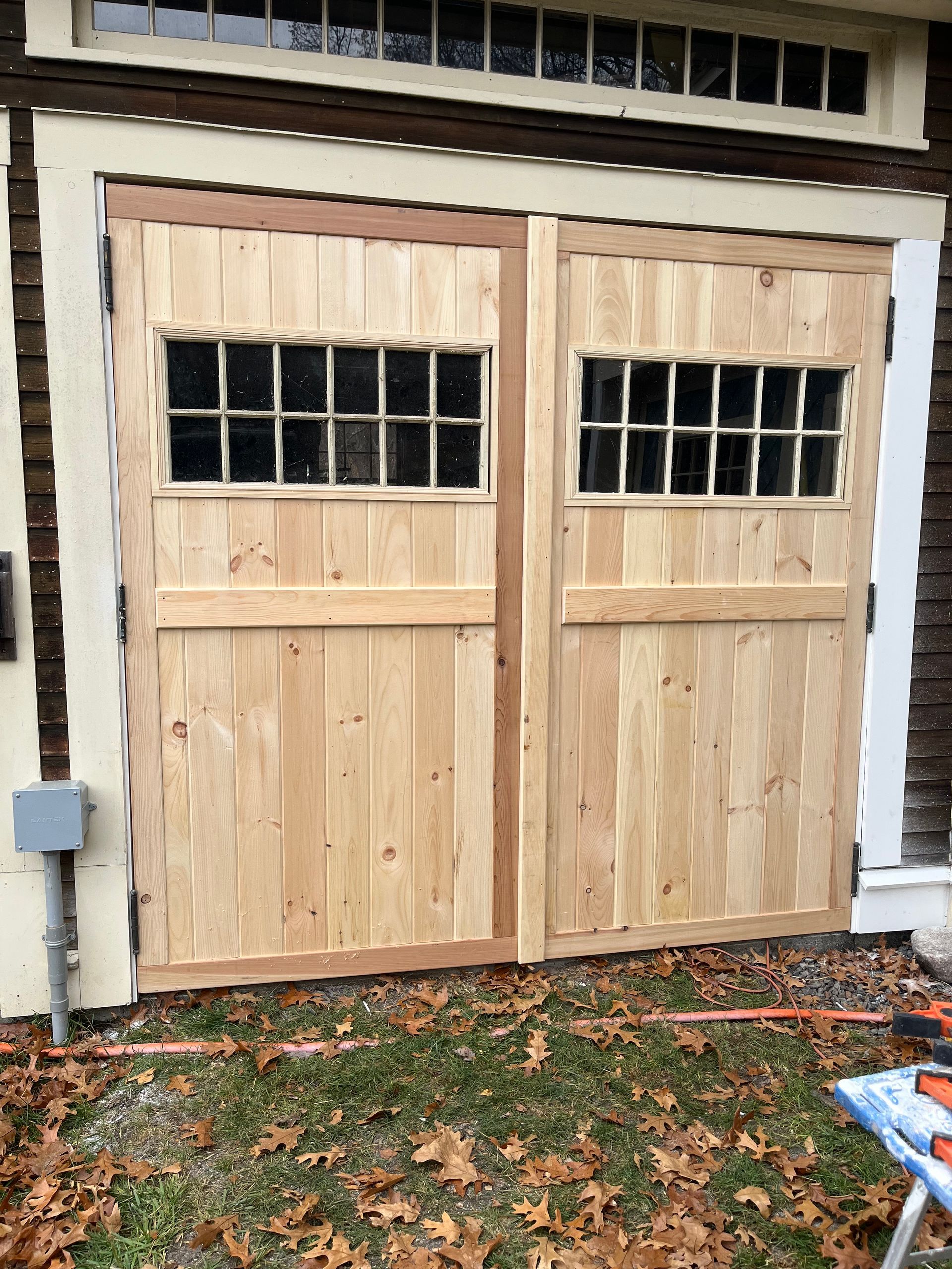 New wooden double garage doors with windows, installed on a rustic building exterior with dry leaves on the ground.