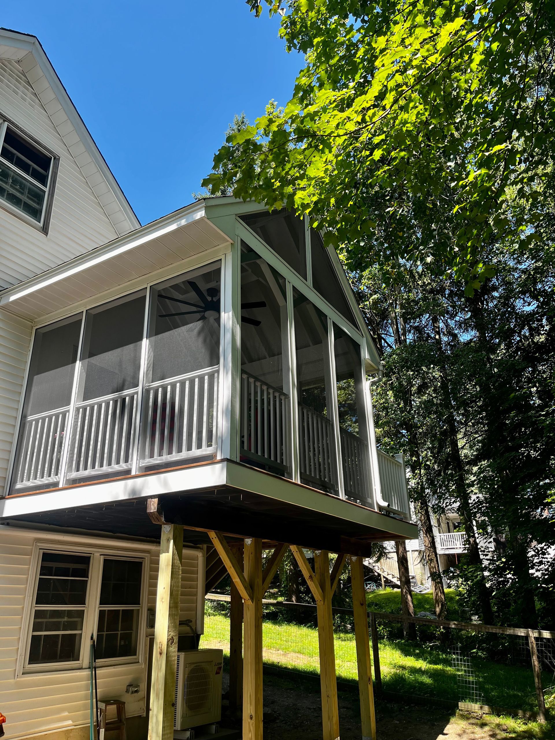 A white house features an elevated, screened-in porch supported by wooden posts above a grassy yard with trees.