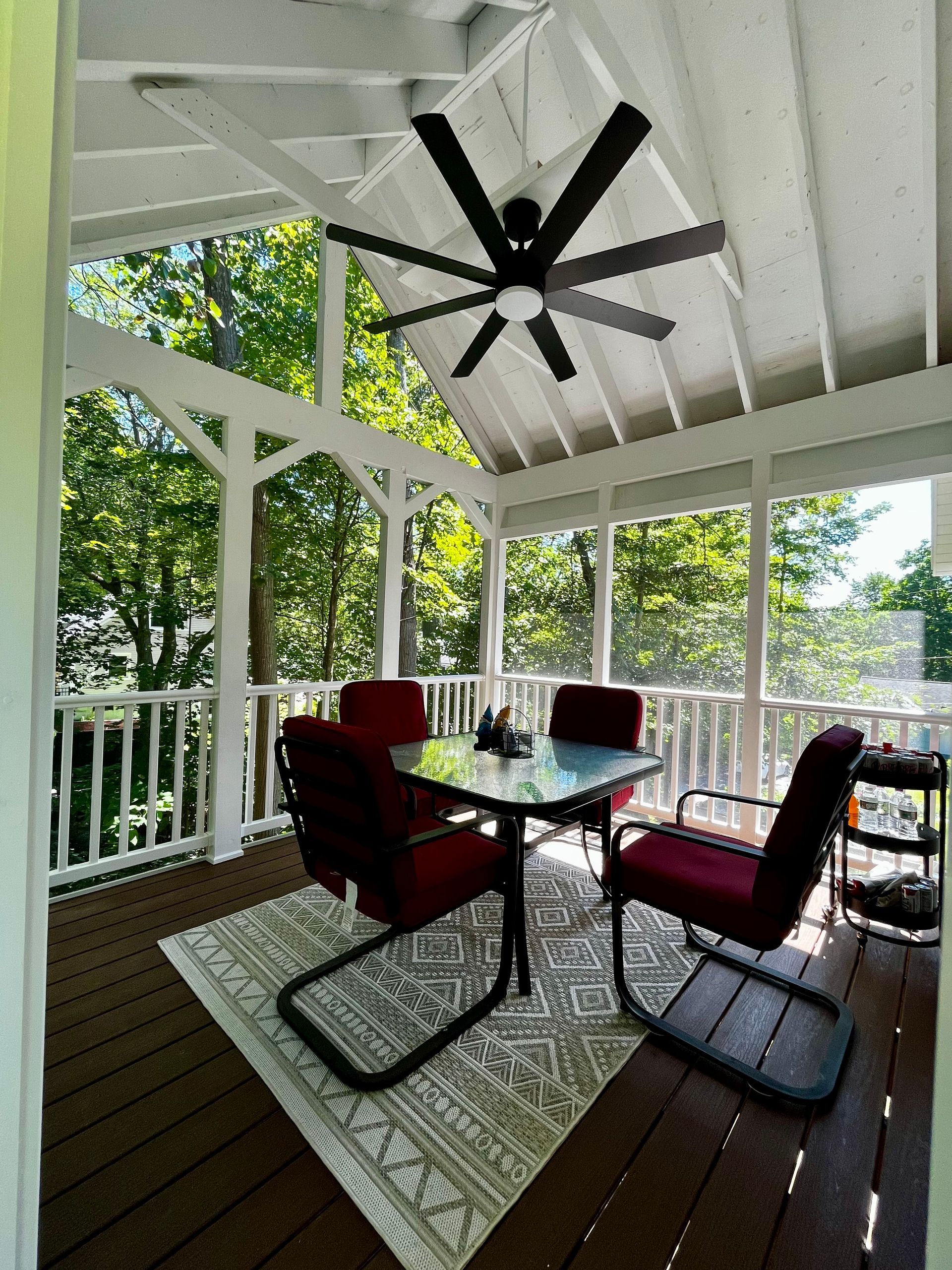 A screened-in deck features a small glass-top table with four burgundy chairs, a patterned area rug, and a ceiling fan.
