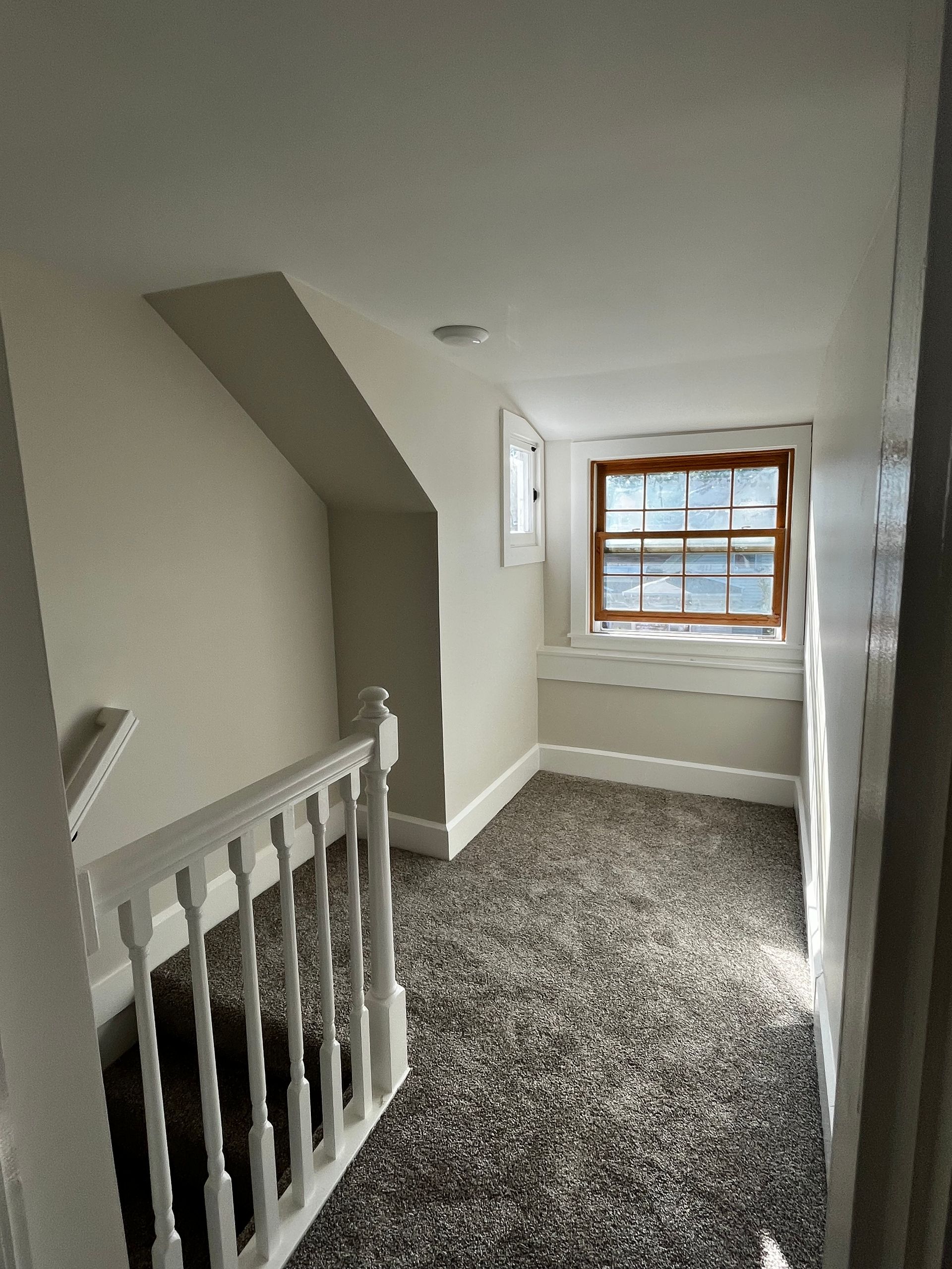 A carpeted landing area with white railings and walls, featuring a wooden window overlooking a brightly lit space.
