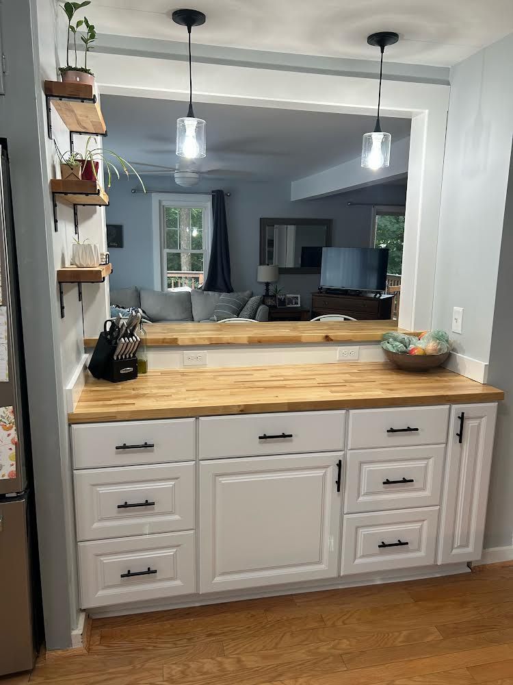 A white kitchen cabinet base with a wooden countertop below a wide wall opening, two pendant lights, and wall shelves.