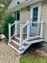 A white-railed, brown wooden front deck and stairs leading to a doorway on a house with beige siding.