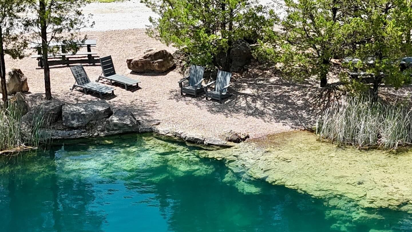 Clear turquoise spring with rocky shore, lounge chairs, and green trees in a natural setting
