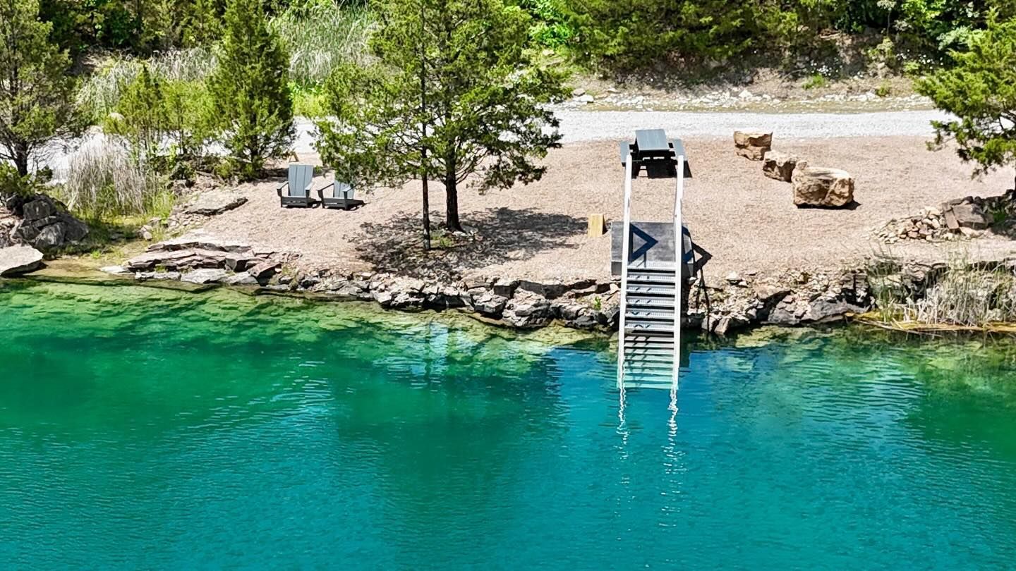 Lakeside dock with a ladder into turquoise water, shaded by trees and lined with picnic tables.