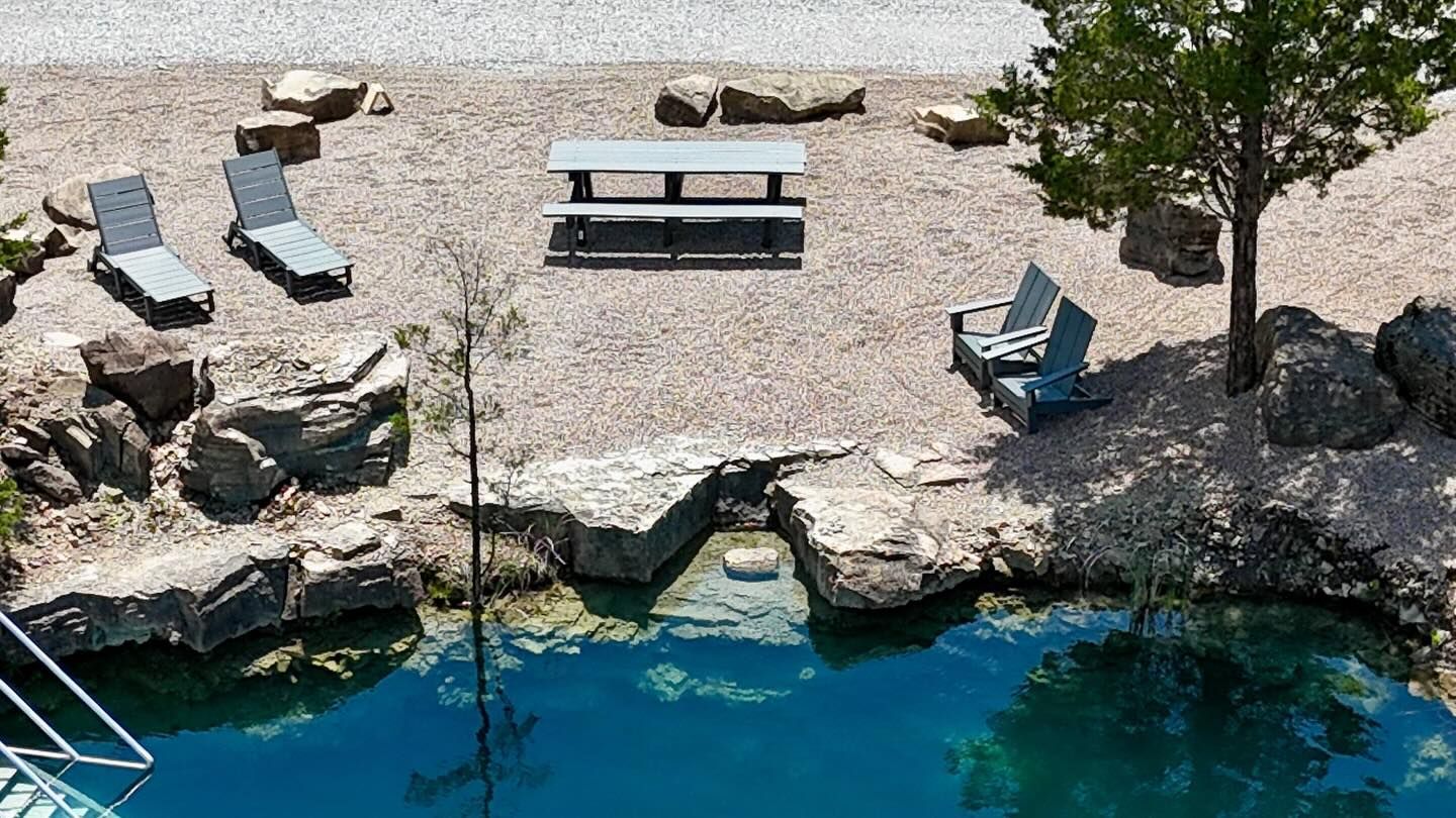 Rocky seaside cove with lounge chairs, picnic table, and a tree beside clear blue water.